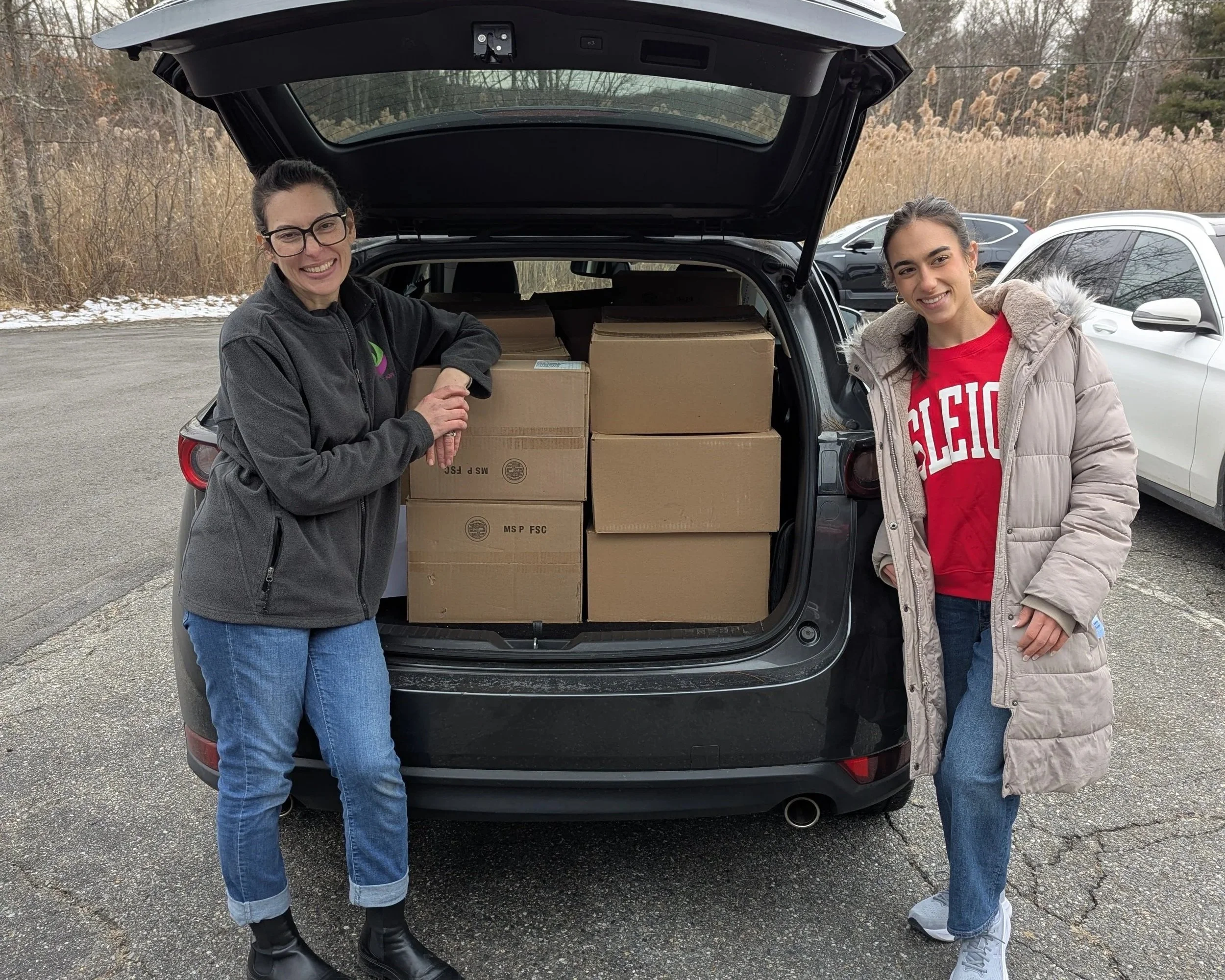 Ari Barbanell and Samantha Asprelli of Give n' Glow stand in front of an open car trunk full of boxes of cosmetics.