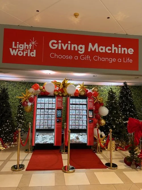 An image of two red vending machines with a festive red and gold balloon arch over them and red carpets leading up to them. There is a banner hanging over them that says "Light the World Giving Machine -- Choose a Gift, Change a Life."
