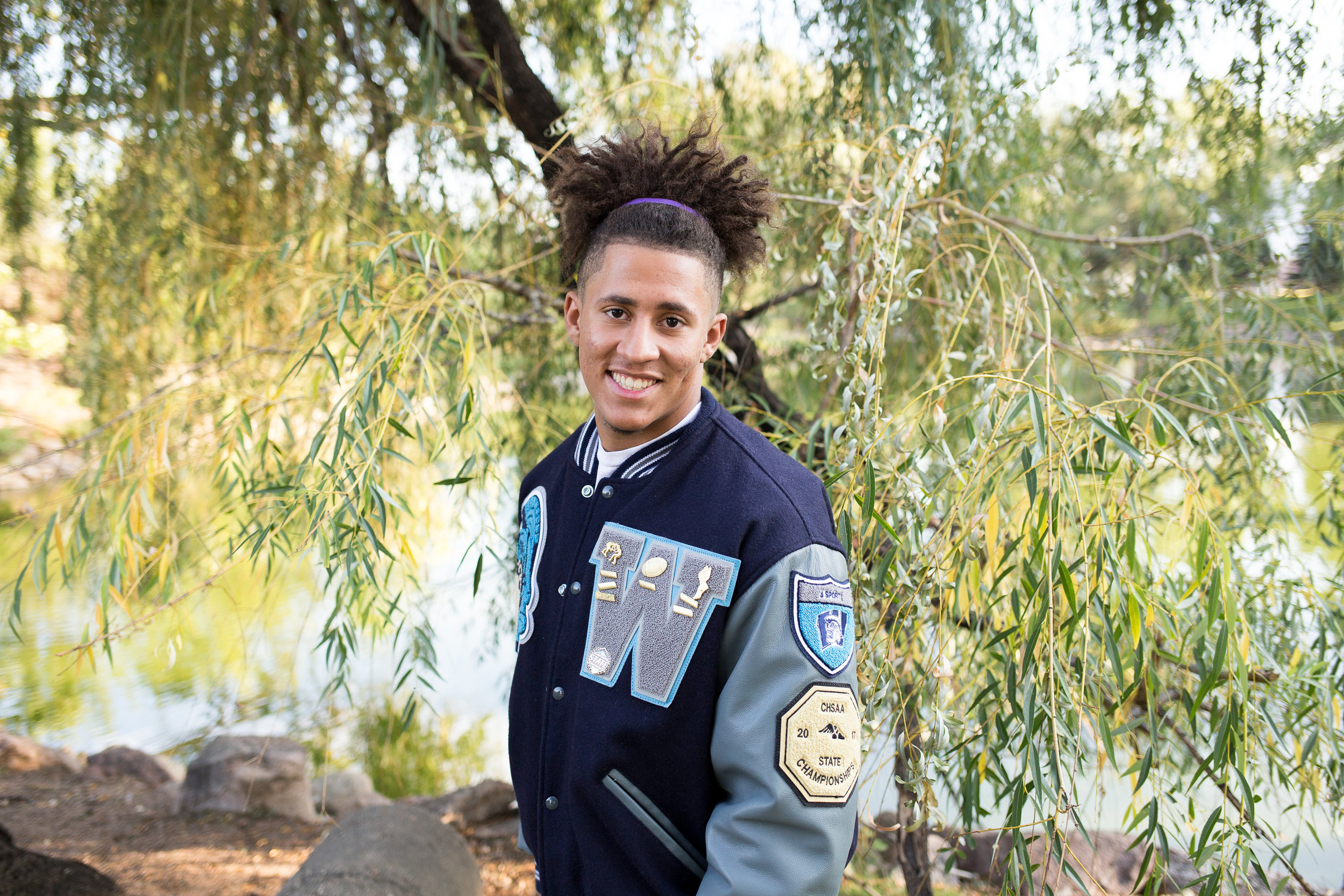 Male senior leaning toward camera in his varsity jacket smiling at camera. Widefield High  School Stacy Carosa Photography
