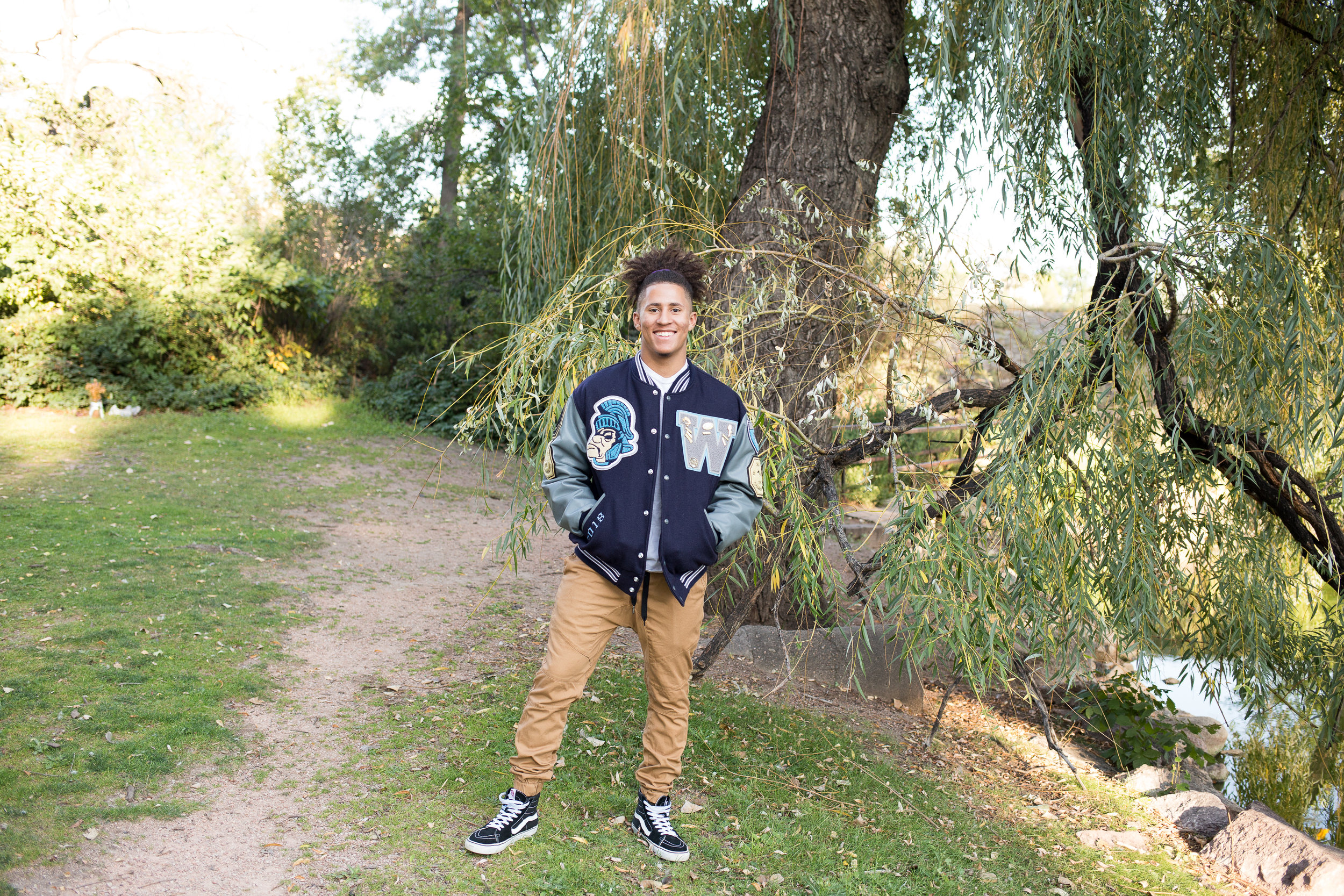 Colorado  Springs Senior Photography Senior standing near trees and pond in Monument Valley Park in his varsity jacket for Widefield High School Stacy Carosa Photography