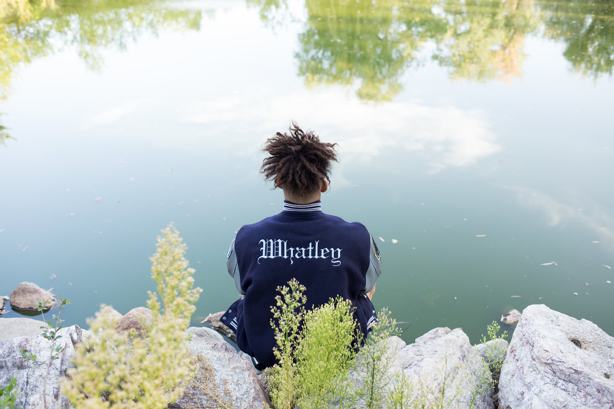 Senior sitting by pond and looking at the water with the back of his varsity jacket facing the camera in Monument Valley Park Stacy Carosa Photography Colorado Springs