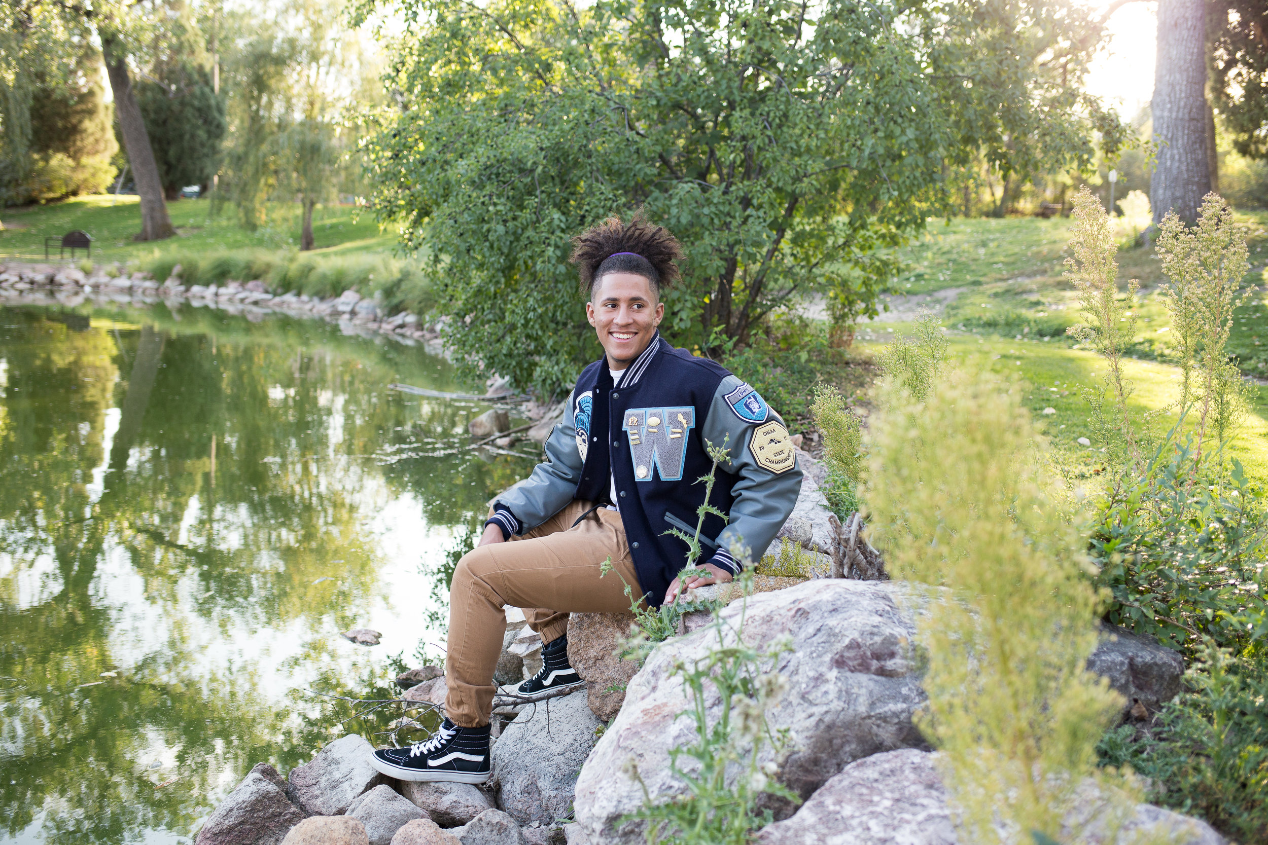 senior sitting on a rock in Monument Valley Park by the pond and turning to look at the camera Colorado Springs senior photography in Colorado Springs Stacy Carosa Photography