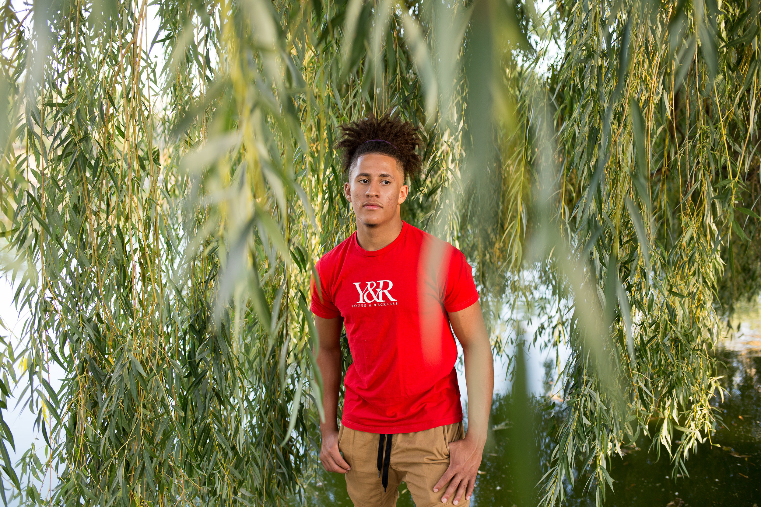 Boy in red shirt standing among the long willow tree branches for his Colorado Springs senior photos in n Monument Valley Park Stacy Carosa Photgraphy