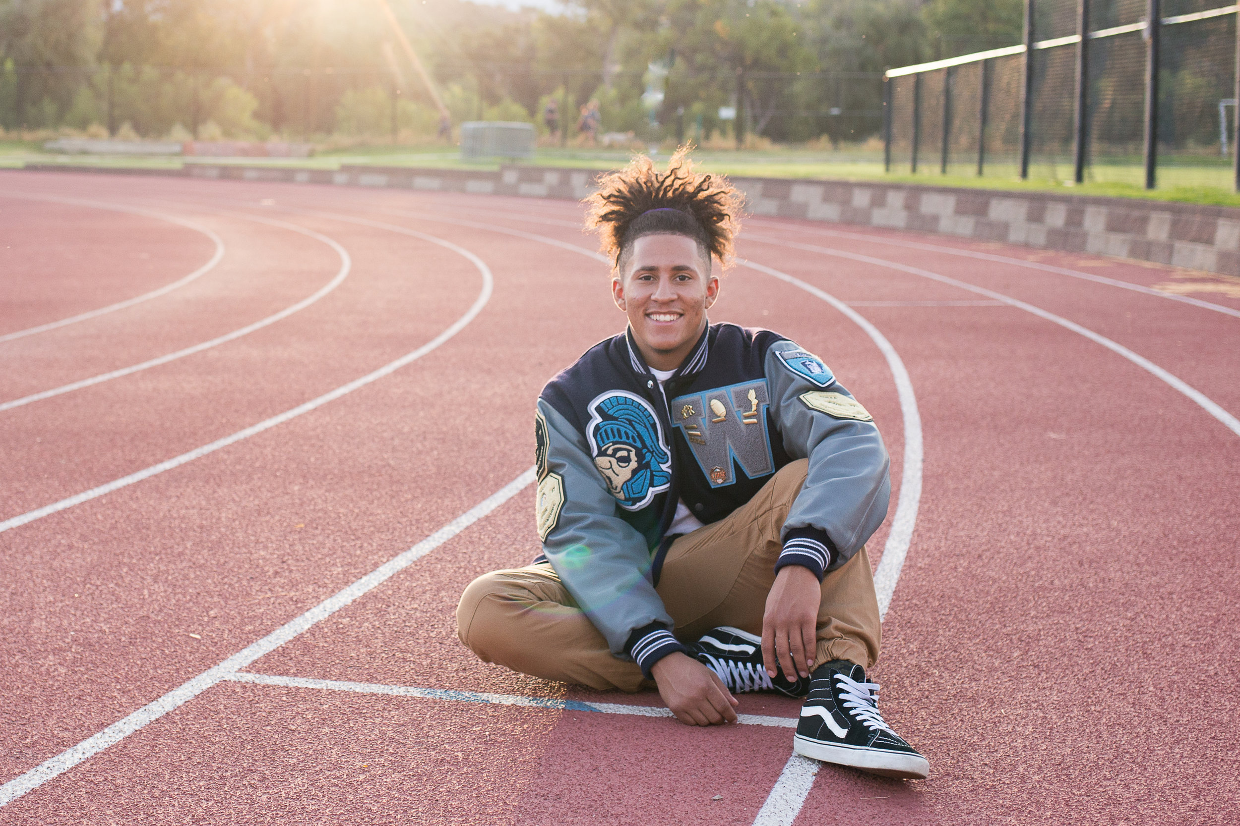 Senior smiling while sitting on a track wearing his varsity jacket and smiling at the camera. Stacy Carosa Photography Colorado Springs senior photography