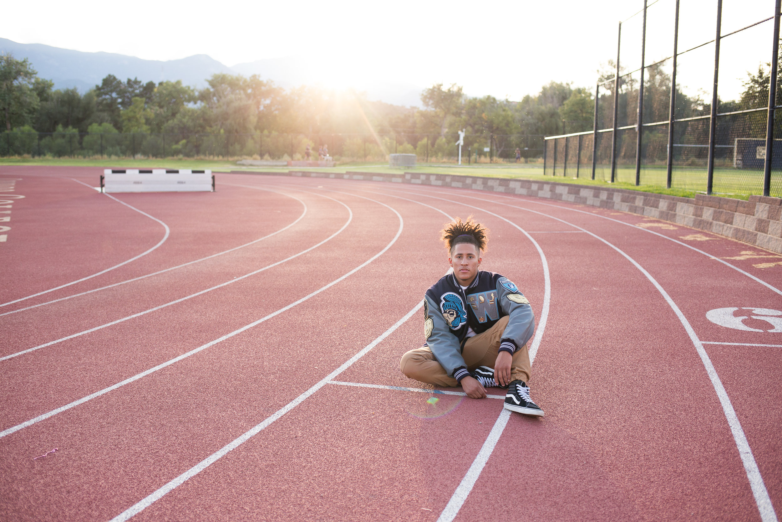 Widefield High School senior in varsity jacket sitting on a track at Colorado College for his Colorado Springs Senior Photos Stacy Carosa Photography