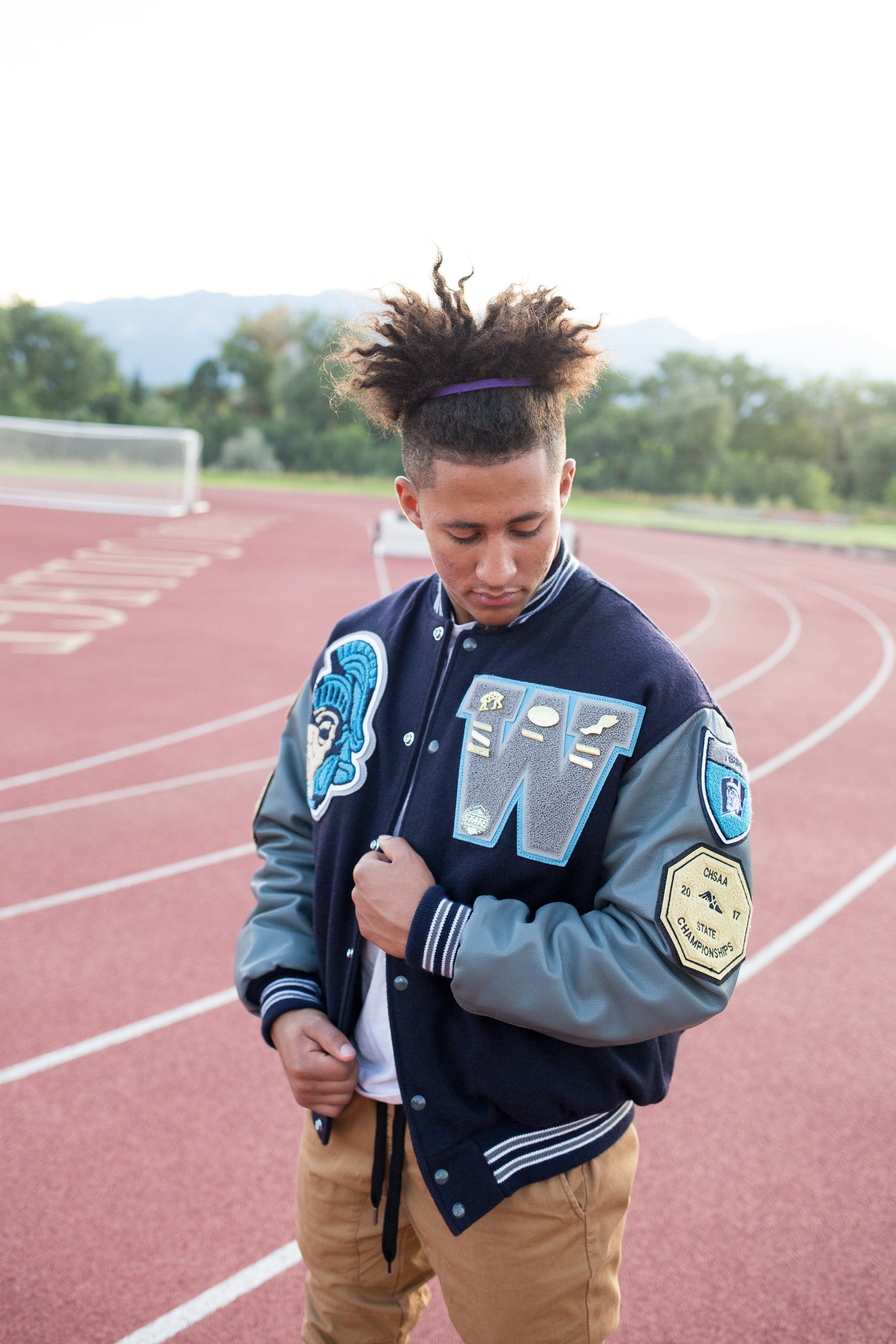 Senior in varsity jacket looking at his badges and pins while standing on a track. Stacy Carosa Photography Colorado Springs senior photography