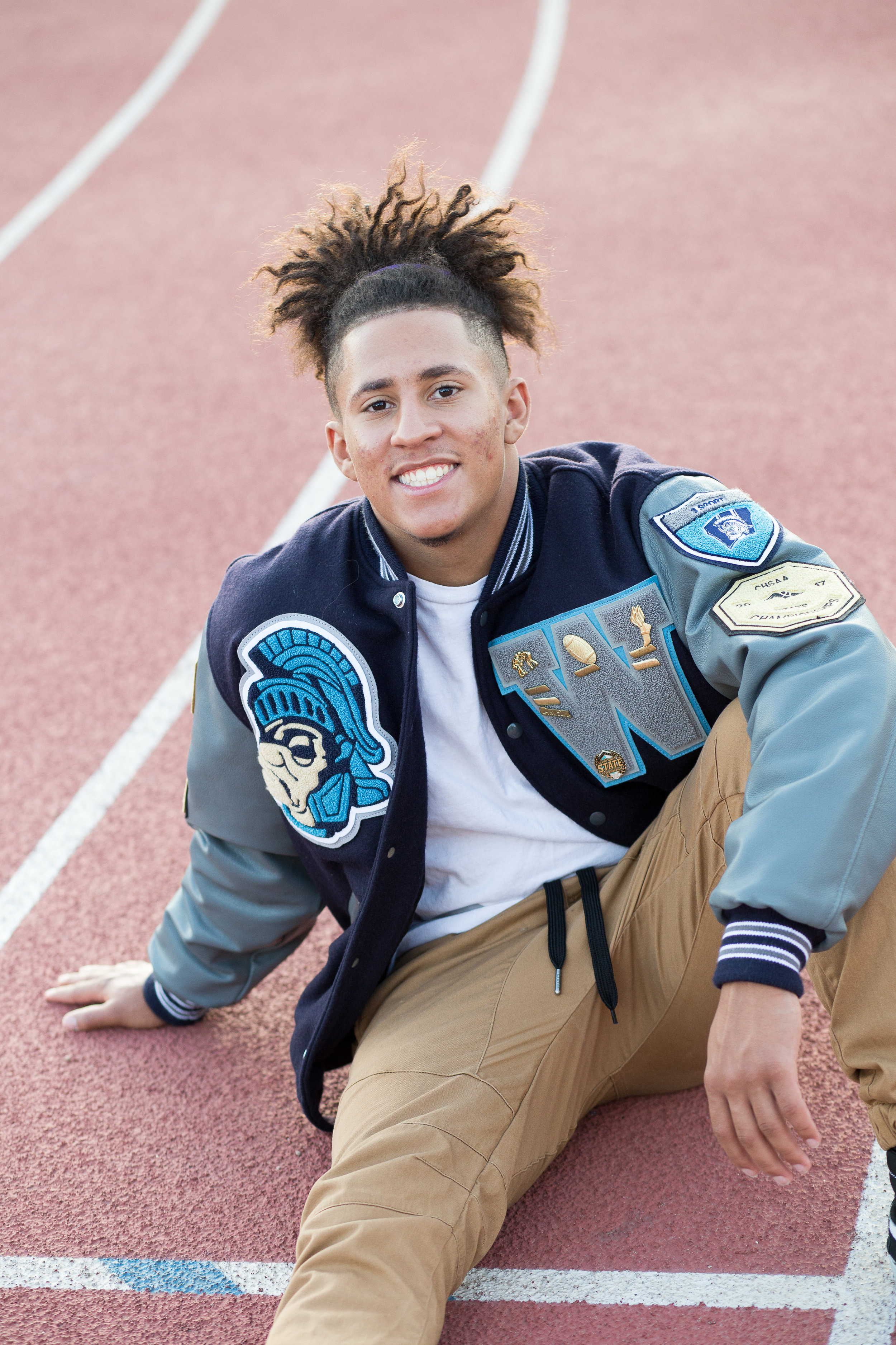 Senior in varsity jacket sitting on a track leaning back on hand smiling at camera Stacy Carosa Photography Colorado Springs