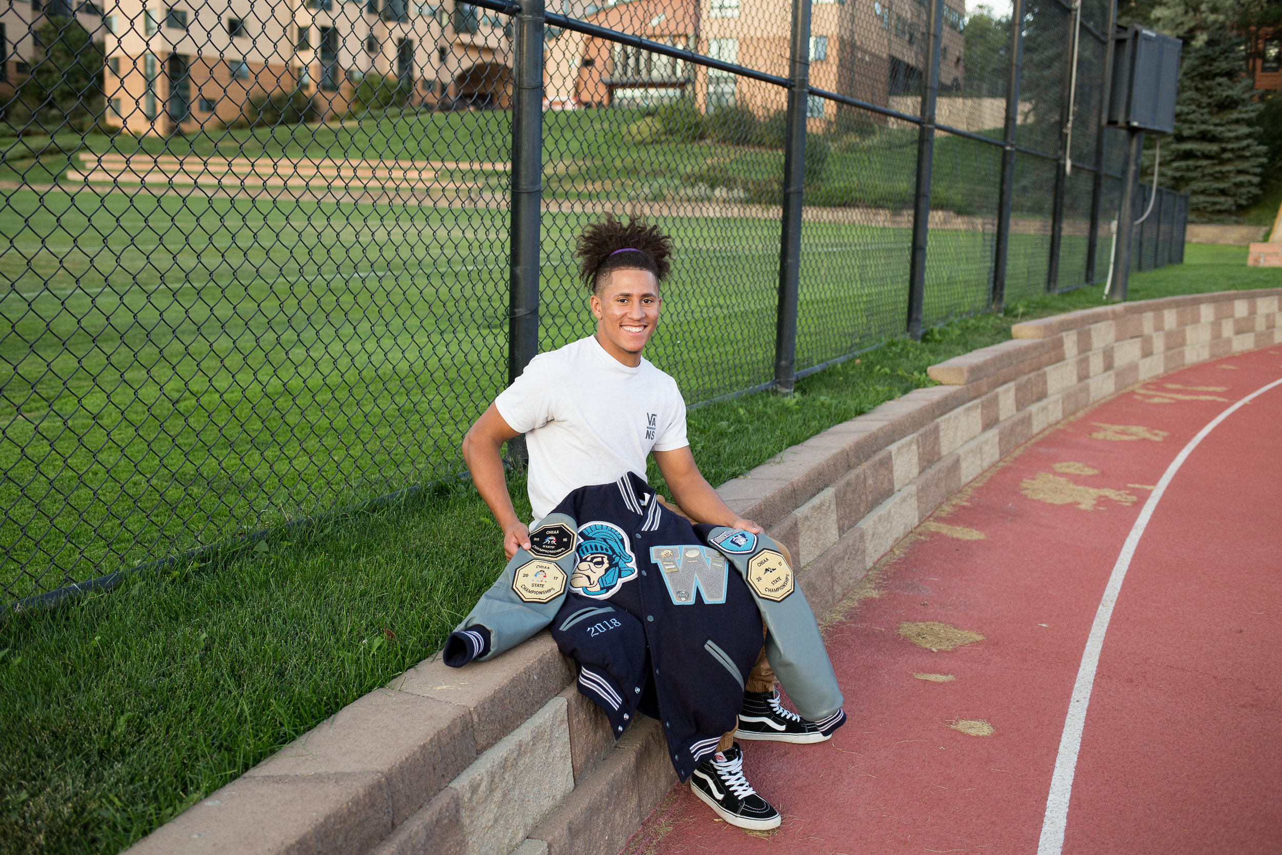 Senior boy sitting on the edge of a track holding his varsity jacket while looking at the camera. Stacy Carosa Photography Colorado Springs Senior Photography