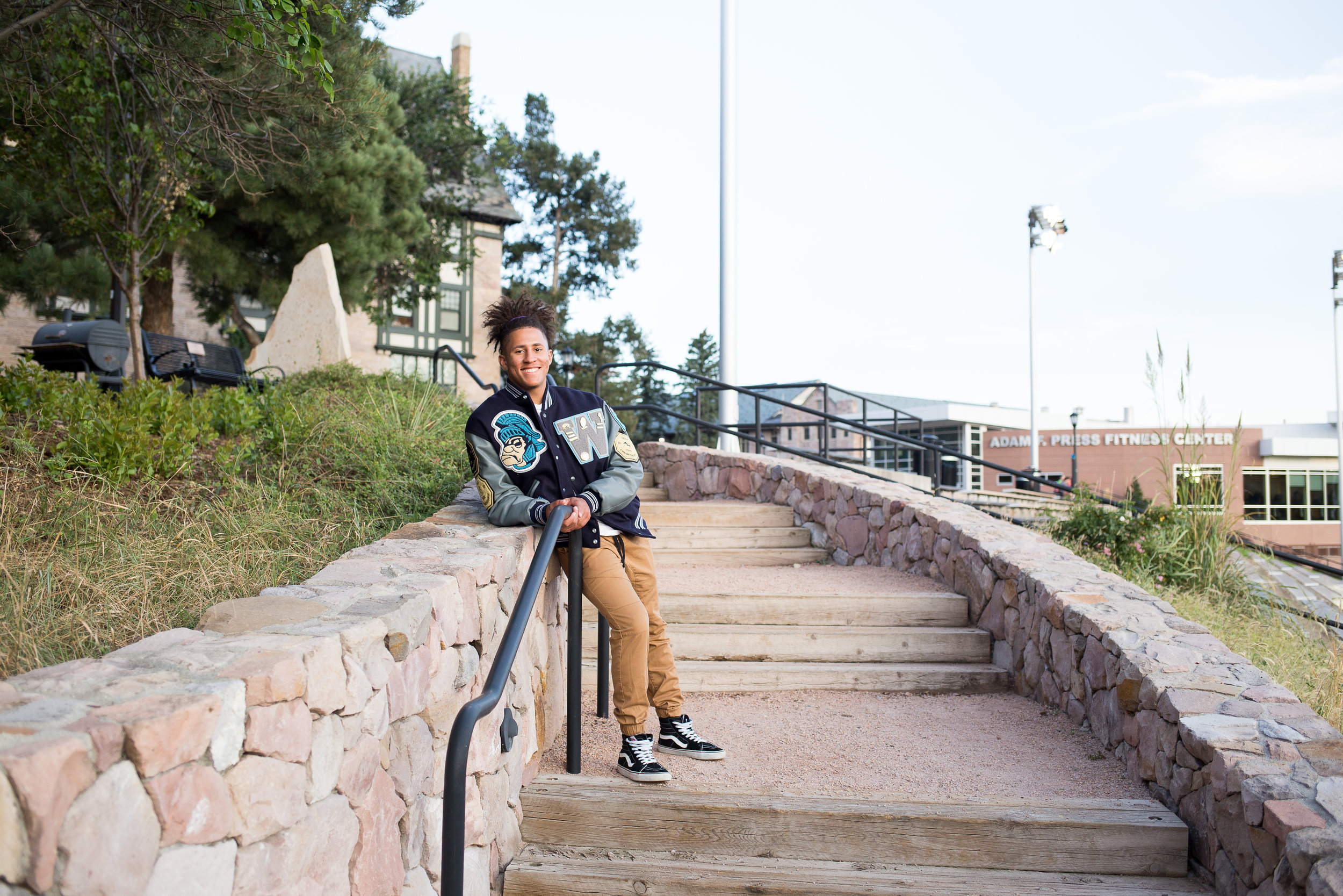 Senior standing on stairs leaning against the stone wall in his varsity jacket Stacy Carosa Photography Colorado Springs senior photos