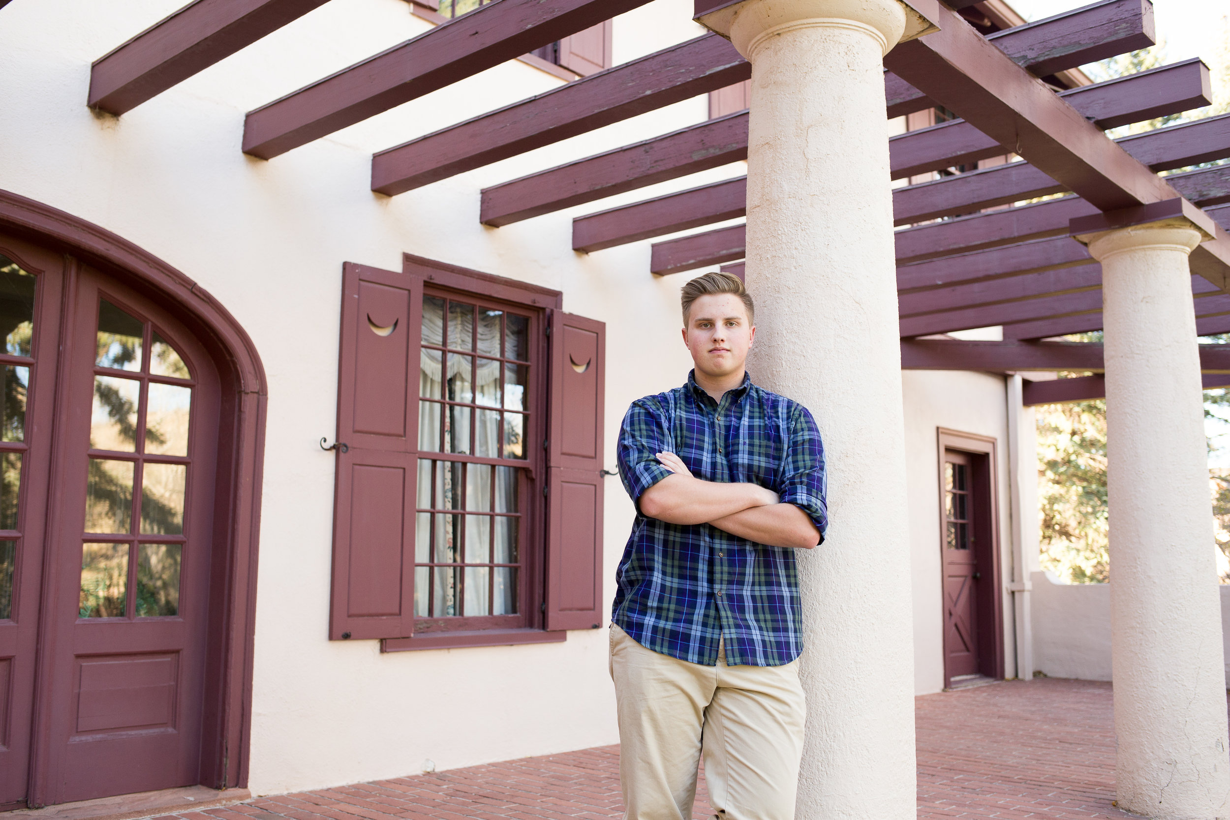 Rock Ledge Ranch Senior Session boy standing and leaning on pillar of a house with arms crossed looking at camera Stacy Carosa Photography