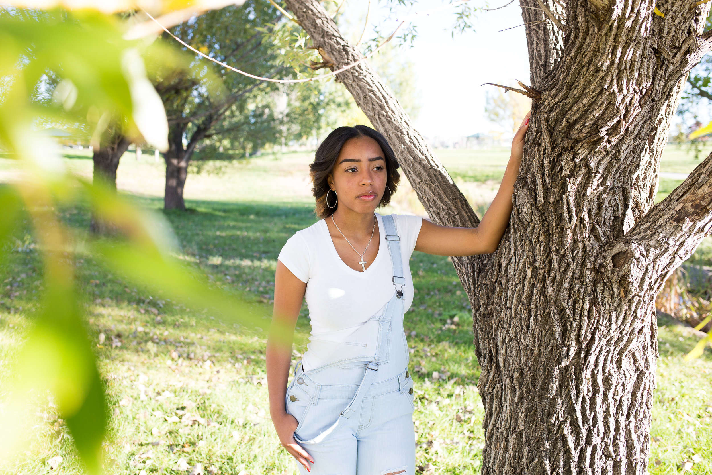 Girl standing near tree with one hand in her overall pockets looking in the distance for her senior photos at Fountain Creek Regional Park in Colorado Springs, CO Stacy Carosa Photography