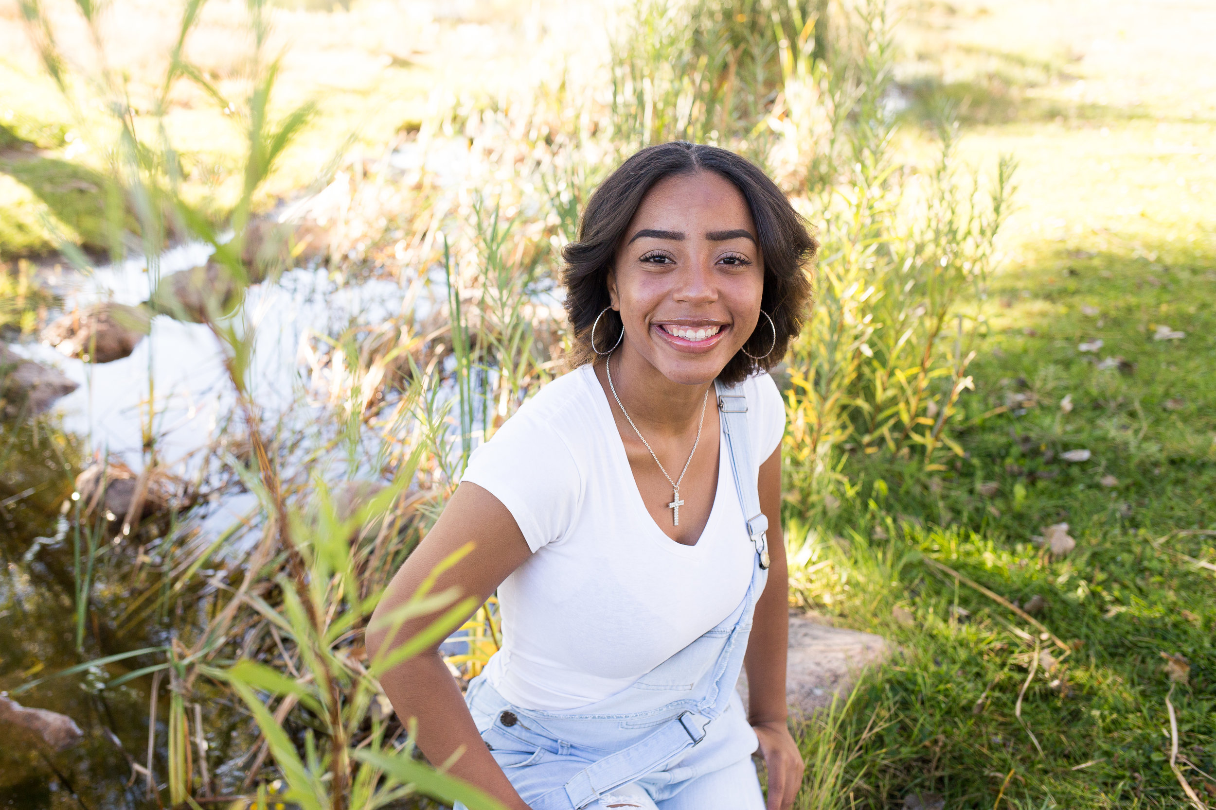 Girl sitting by creek and tall grasses smiling at camera for her senior photos in Fountain Creek Regional Park Widefield High School Stacy Carosa Photography Senior Photos