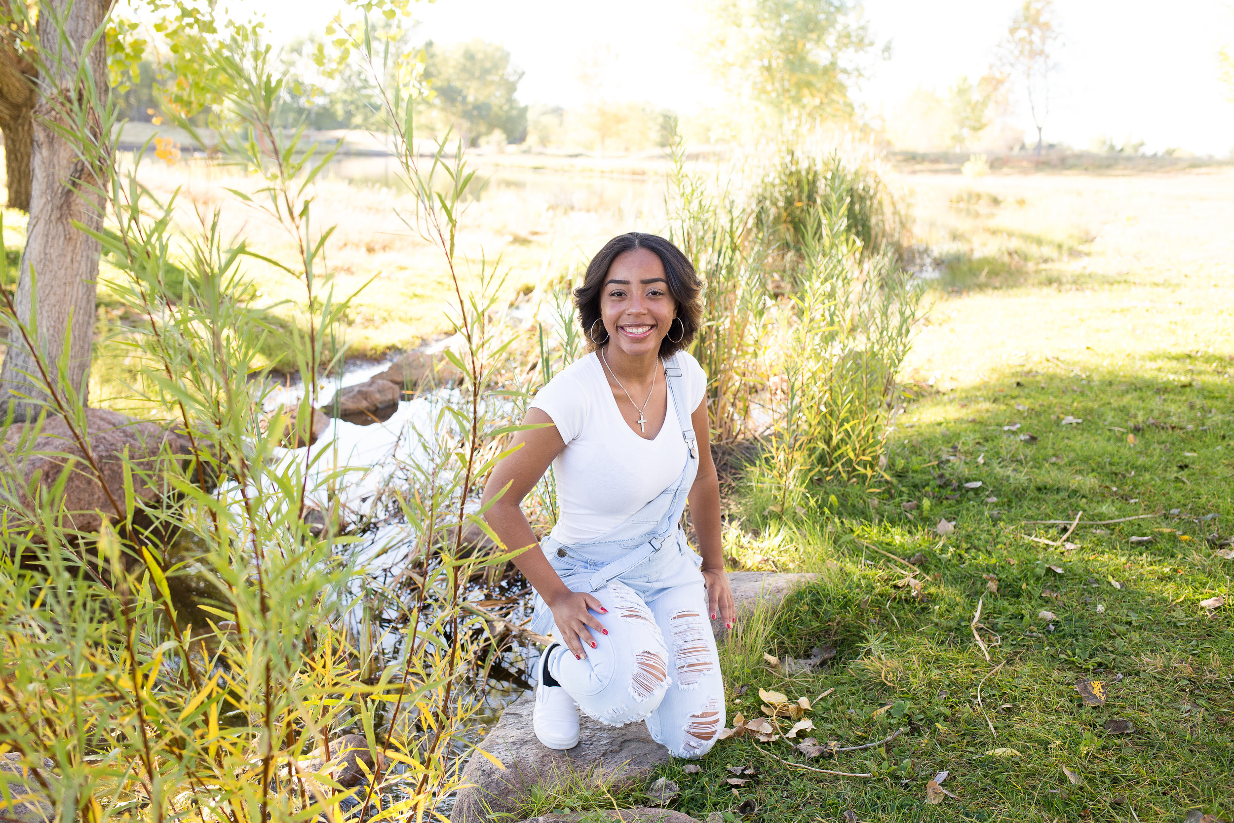 Girl kneeling by creek and tall grasses in overalls at Fountain Creek Regional Park Colorado Springs Senior Portraits Stacy Carosa Photography
