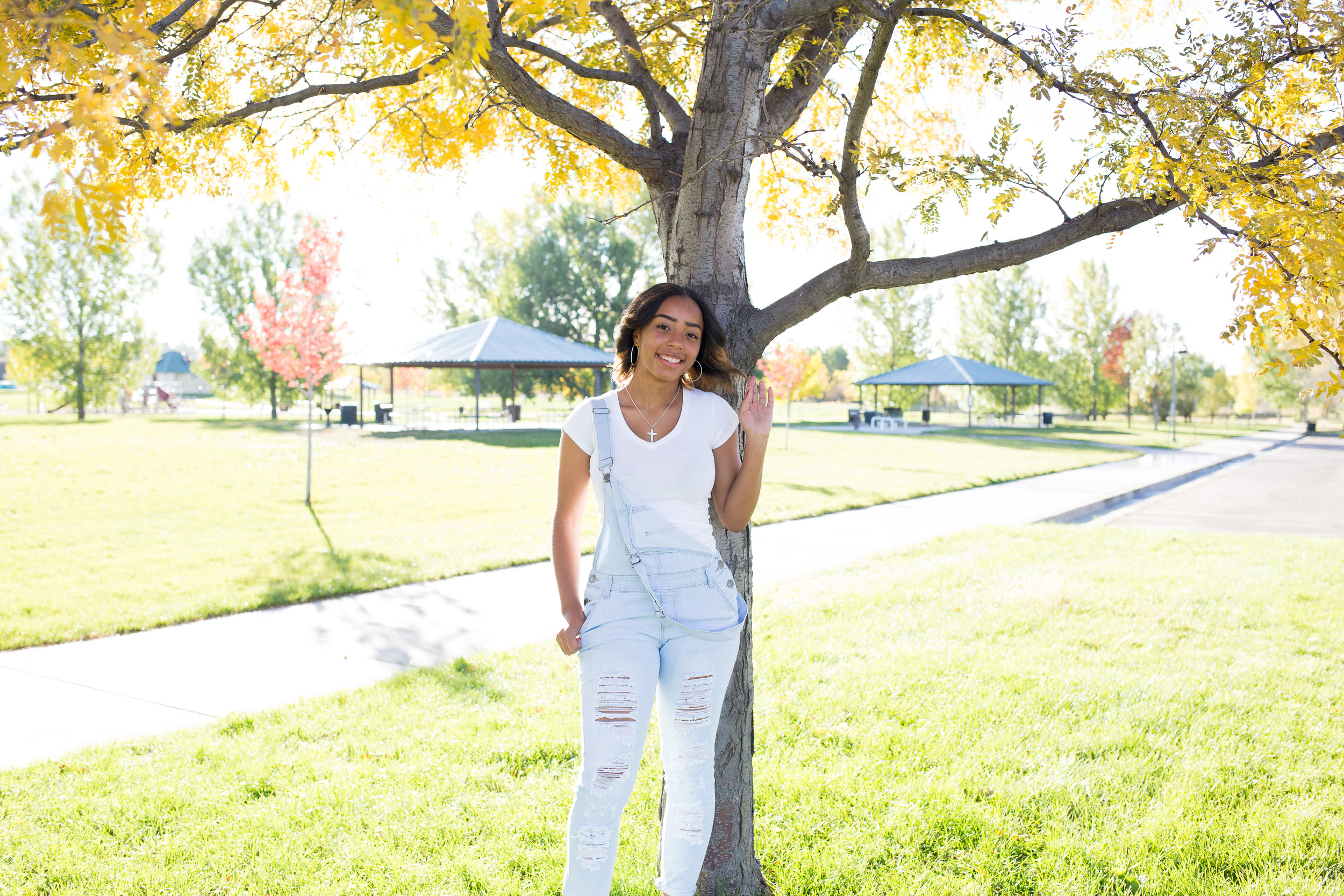 Widefield High School girl in overalls leaning again a tree with fall colors for her senior photos at Fountain Creek Regional Park in Colorado Springs, CO Stacy Carosa Photography