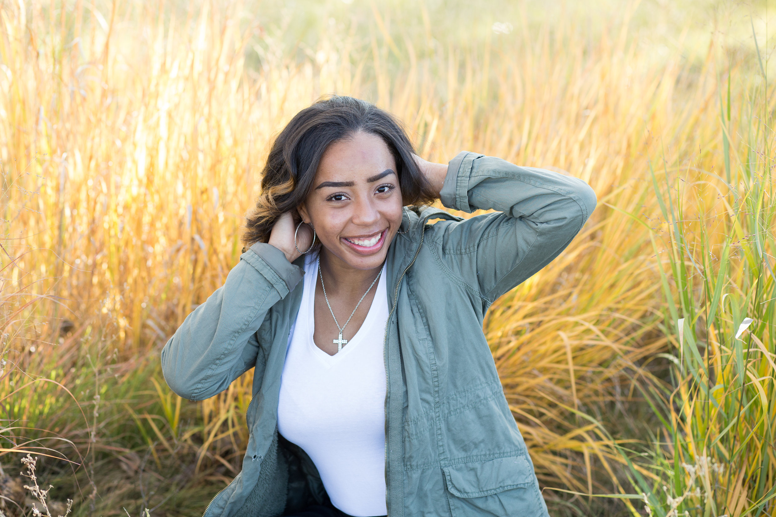 Senior girl sitting in the tall grasses smiling at camera for her senior session at Fountain  Creek Regional Park Stacy Carosa Photography Colorado Springs Senior Photographer
