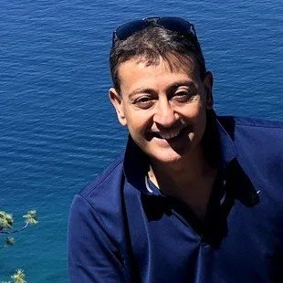 A smiling man with short brown hair, wearing sunglasses and a dark blue shirt near a large body of water.