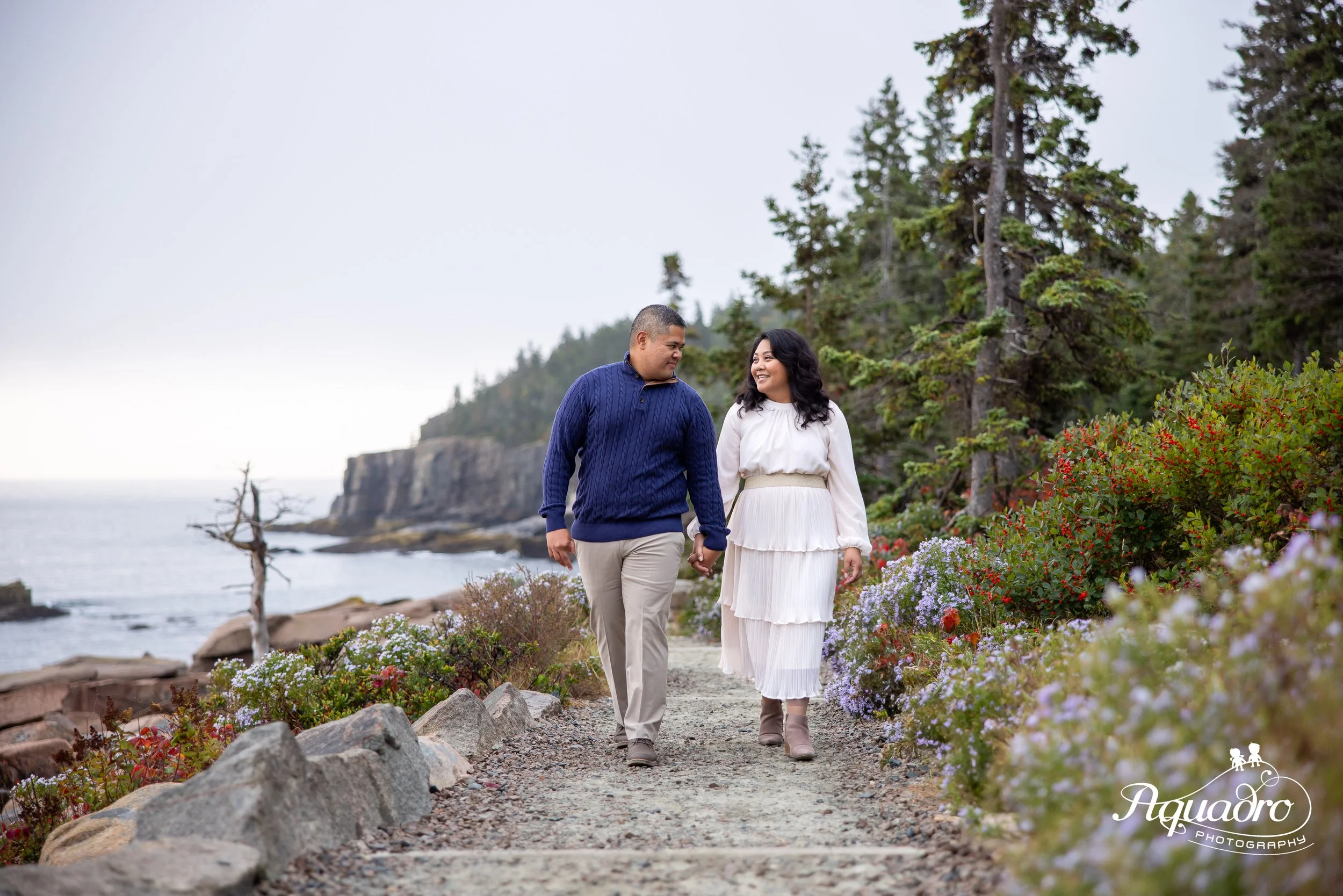 couple walks along ocean path with otter cliffs and wildflowers in acadia national park on mount desert island near bar harbor