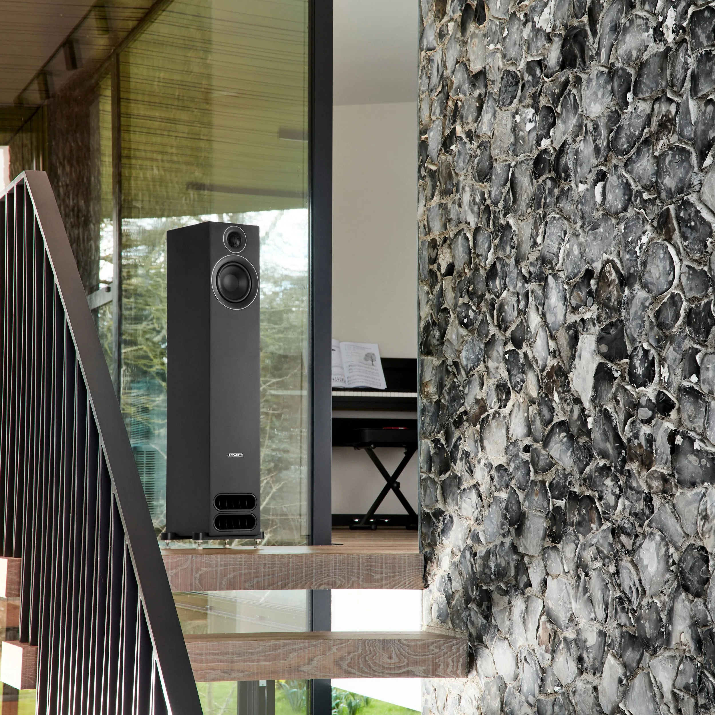 A PMC prodigy5 floorstanding speaker pictured in front of glass and beside a stone wall.