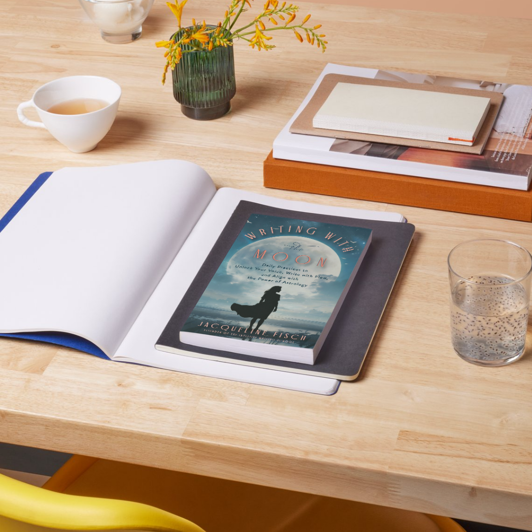 Writing with the moon book by Jacqueline Fisch sitting on a light wood desk with notebooks, coffee, and water