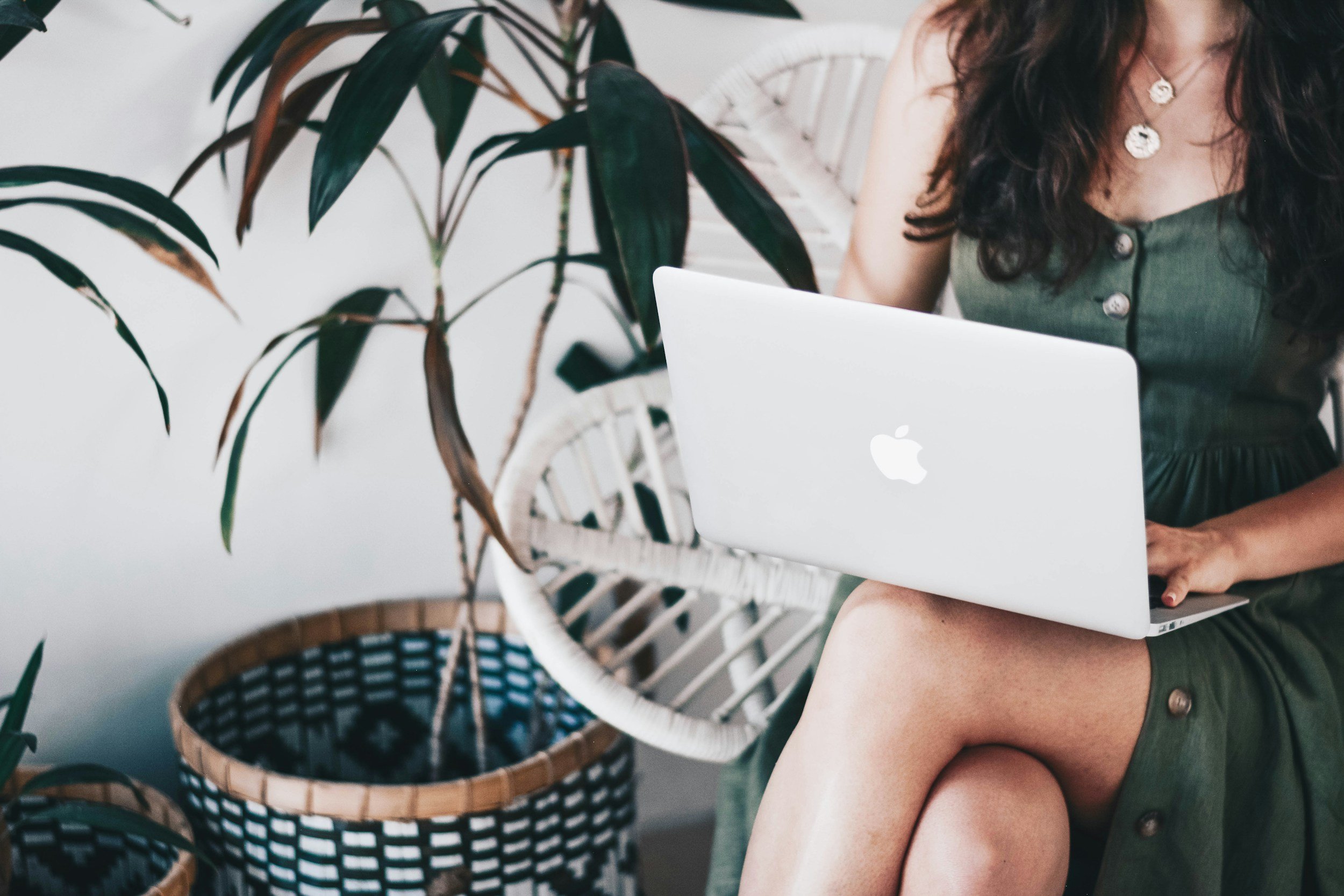woman in a green dress with dark hair writing on a macbook sitting in a wicker chair next to a plant
