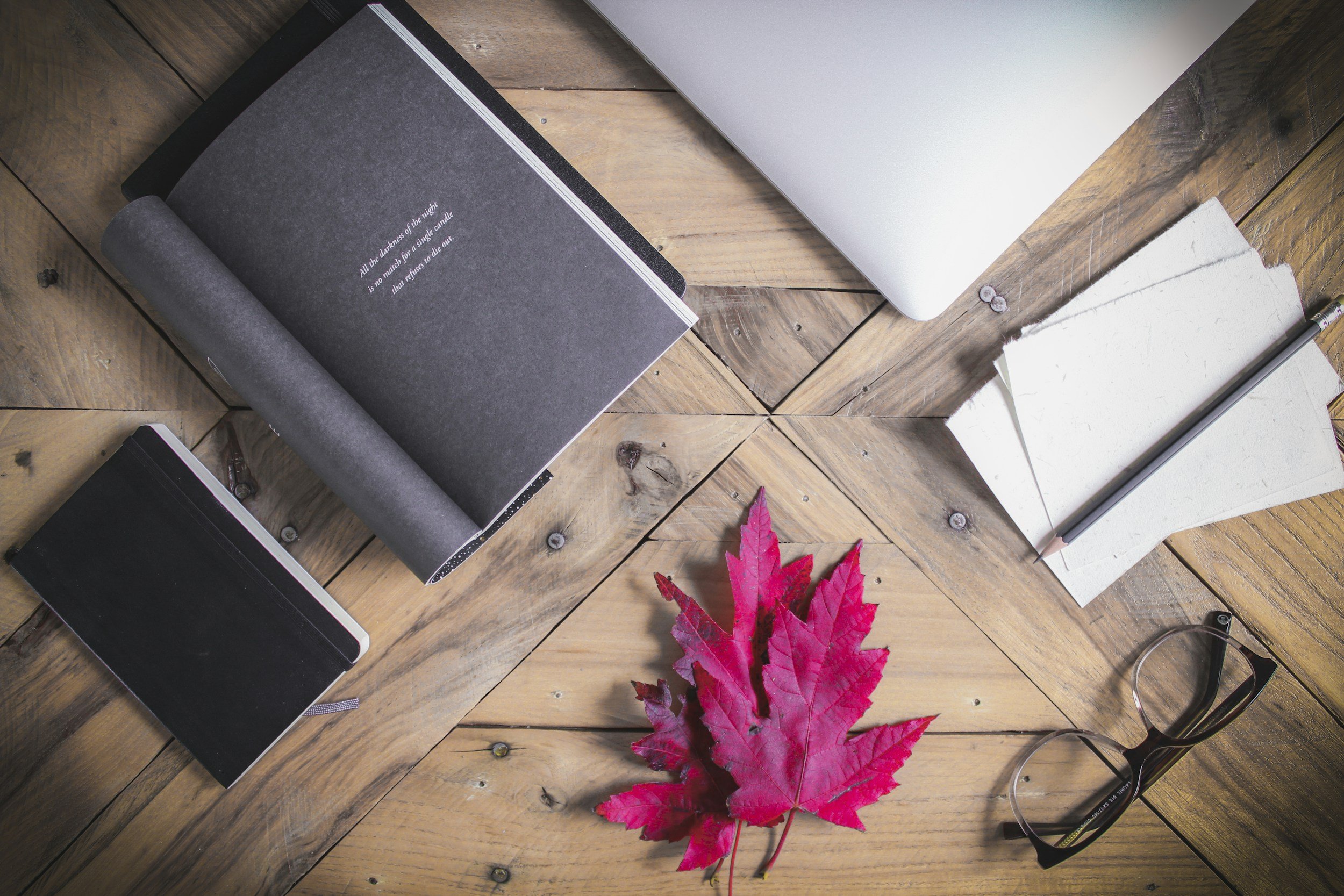 A red leaf, laptop, index cards, and a notebook on a wooden desk