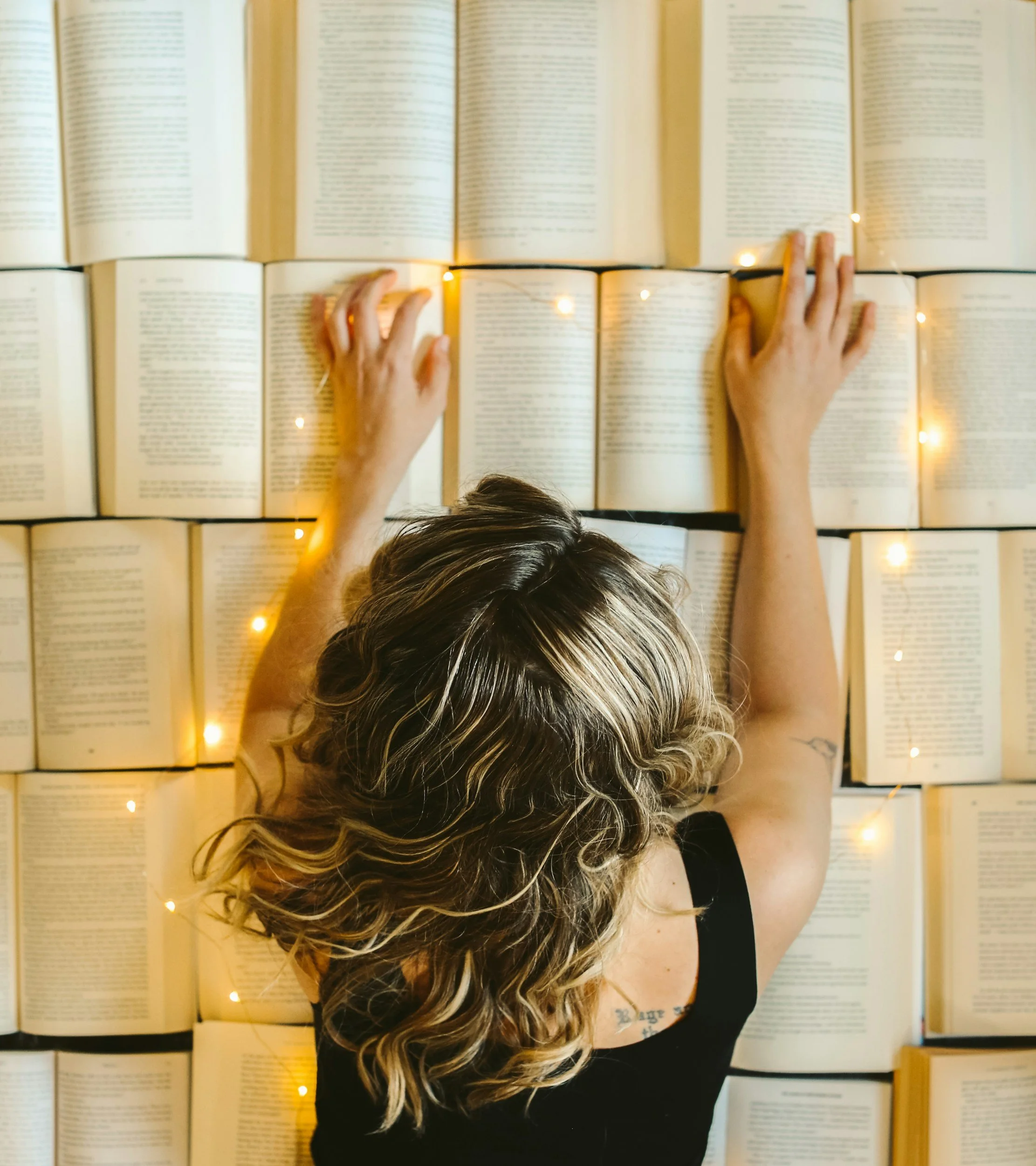 A person with curly brown and blonde hair is standing in front of multiple open books on a wall, reaching out to touch or adjust one of the books. The person's face is not visible, and they are wearing a black shirt with a tattoo on their left arm.