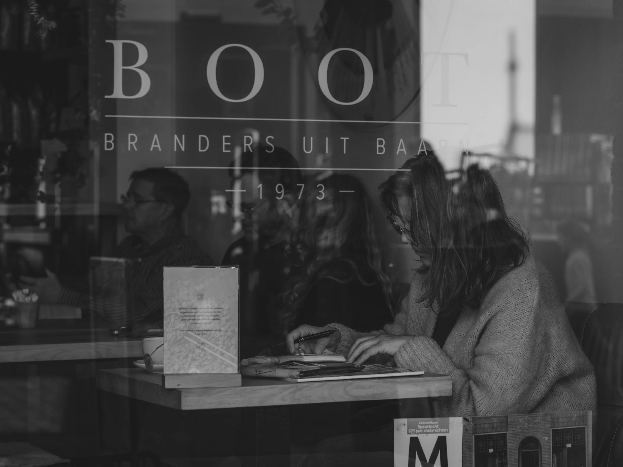 People sitting at a table inside a bookstore café, viewed through the glass window with the store's name 'BOOK' and other text. The individuals are engaged in various activities, possibly reading or using devices.