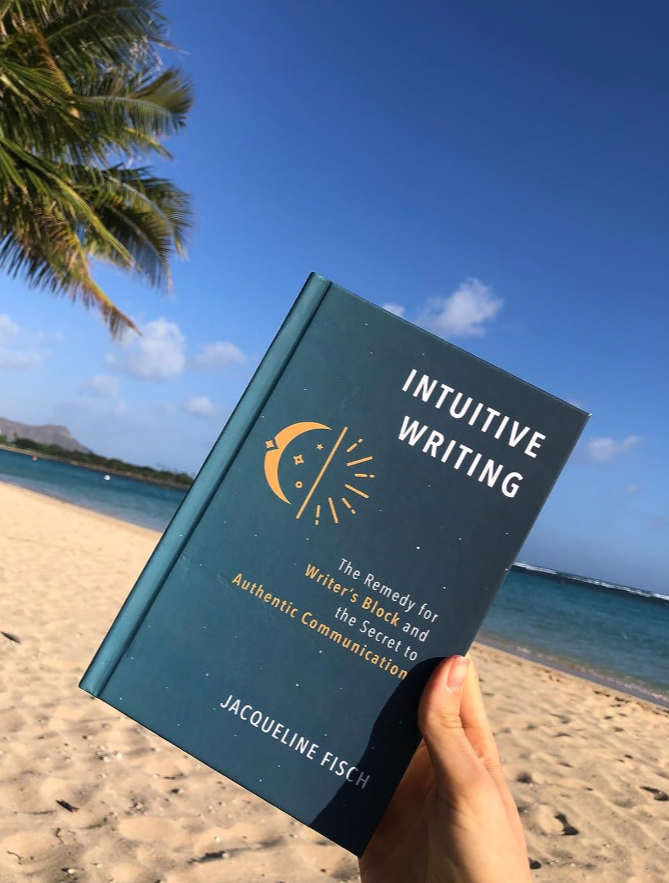 A person holding a book titled "Intuitive Writing" on a beach with sand, palm trees, and calm ocean water under a blue sky with a few clouds.