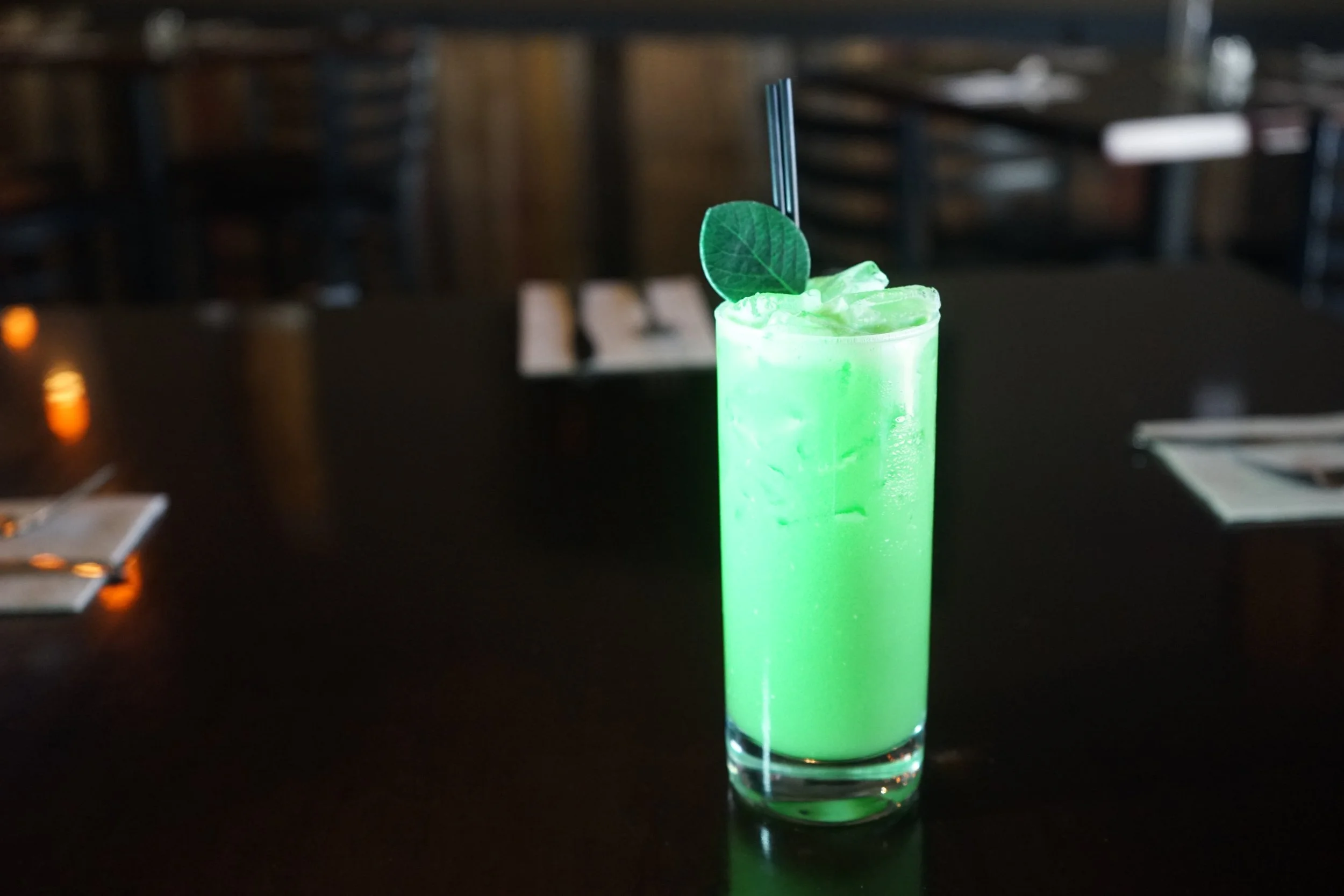 Bright green cocktail in a tall glass with ice, garnished with a leaf and two black straws, on a dark table in a dimly lit restaurant.