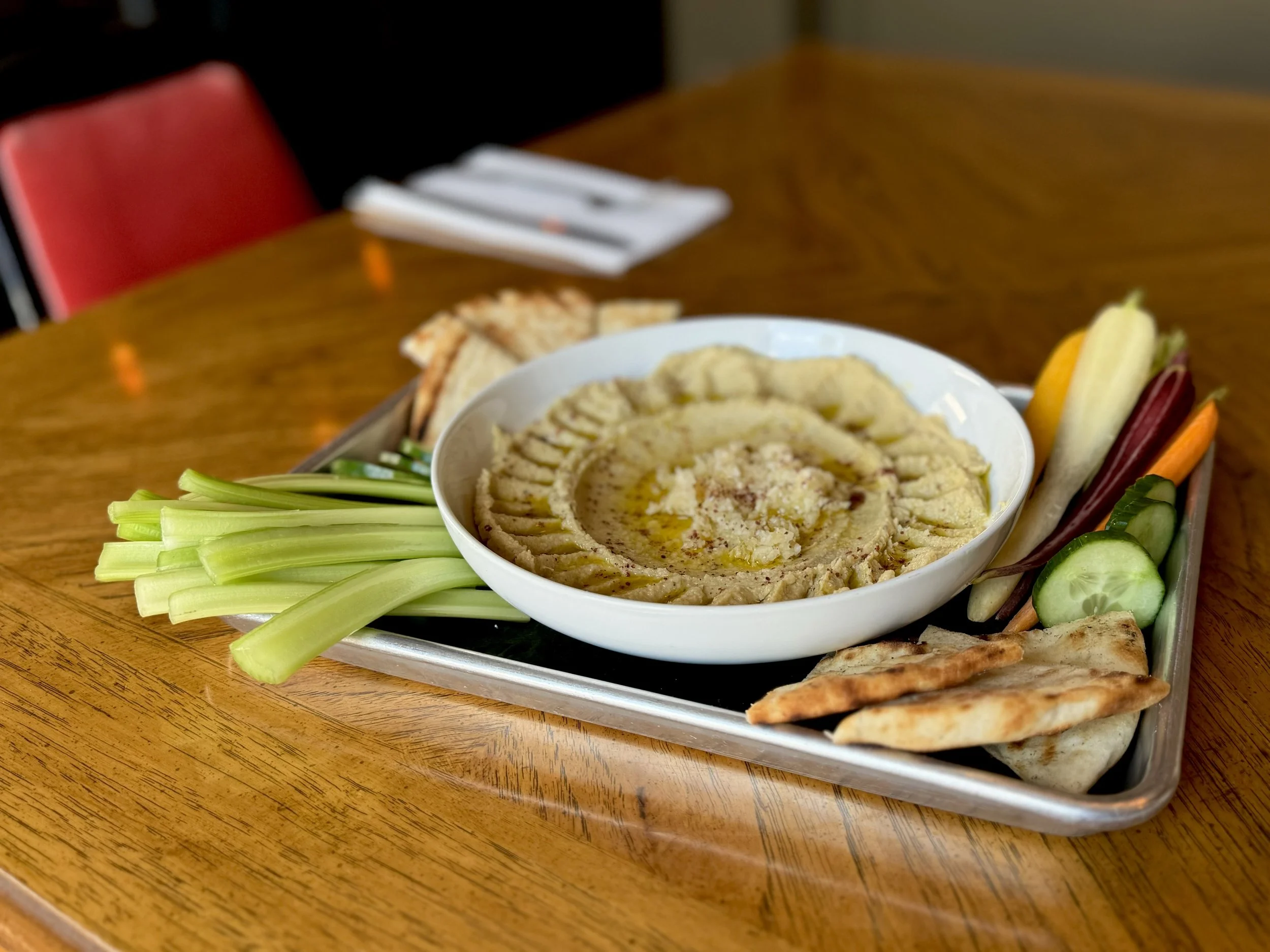 A plate of hummus with olive oil and spices surrounded by pita bread, celery sticks, carrots, and cucumber slices on a wooden table.