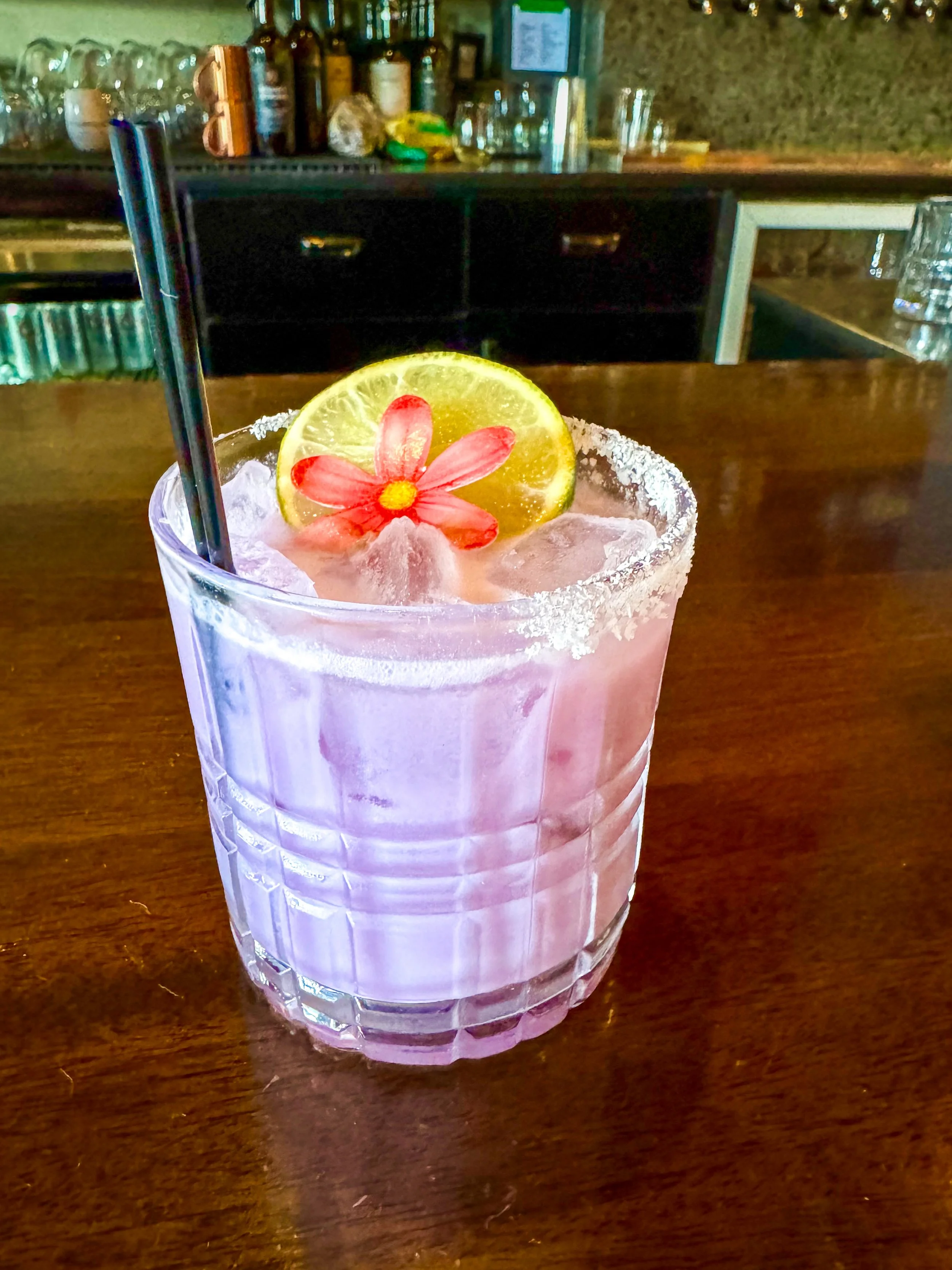 A pink cocktail in a glass with a salted rim, garnished with a lime slice and a pink flower, two black straws on a wooden bar.
