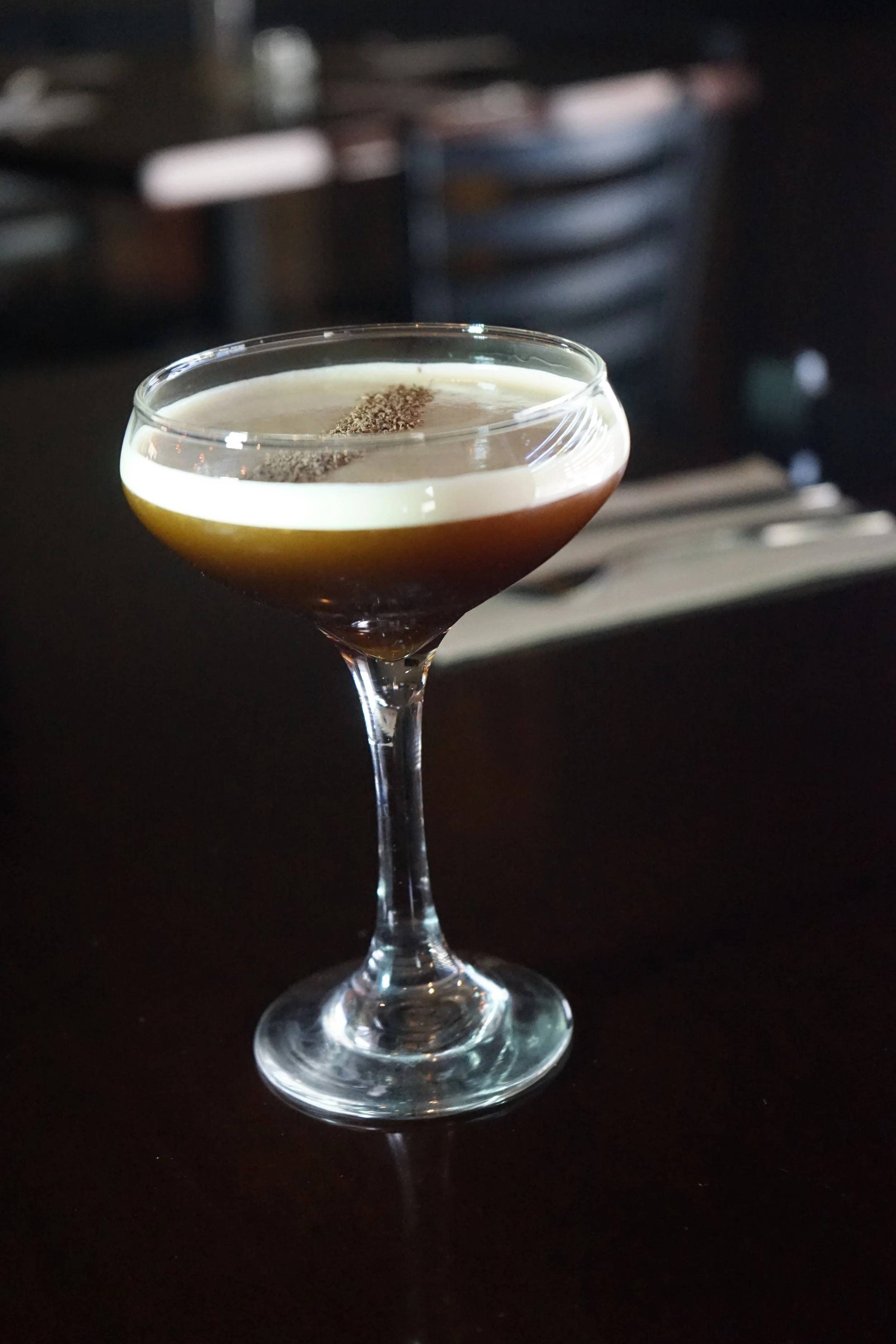 A cocktail served in a coupe glass on a dark surface, featuring a frothy top layer and a sprinkle of ground spices.