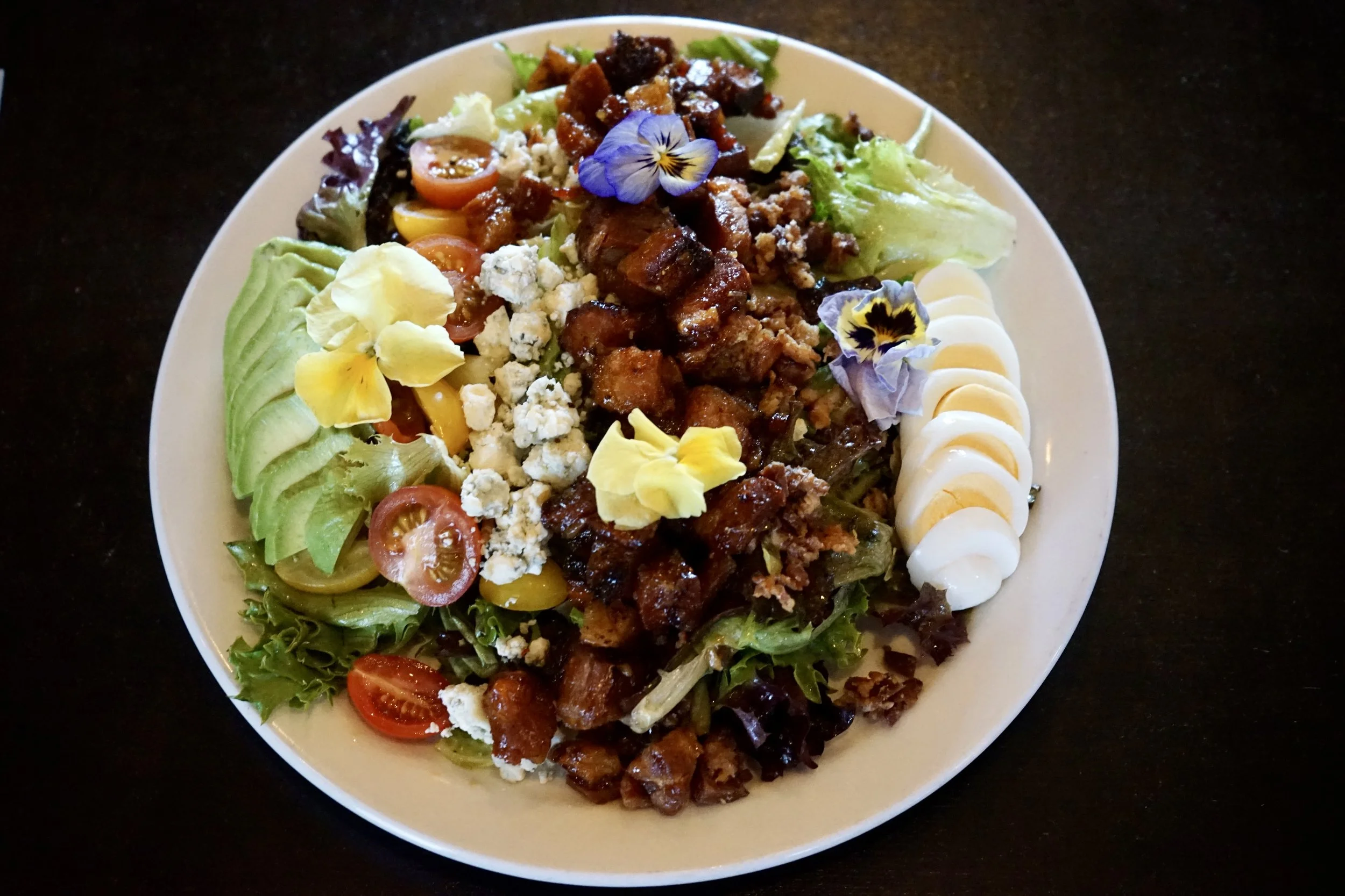 Salad with sliced avocado, cherry tomatoes, crumbled blue cheese, diced meat, hard-boiled egg slices, and edible flowers on a plate.