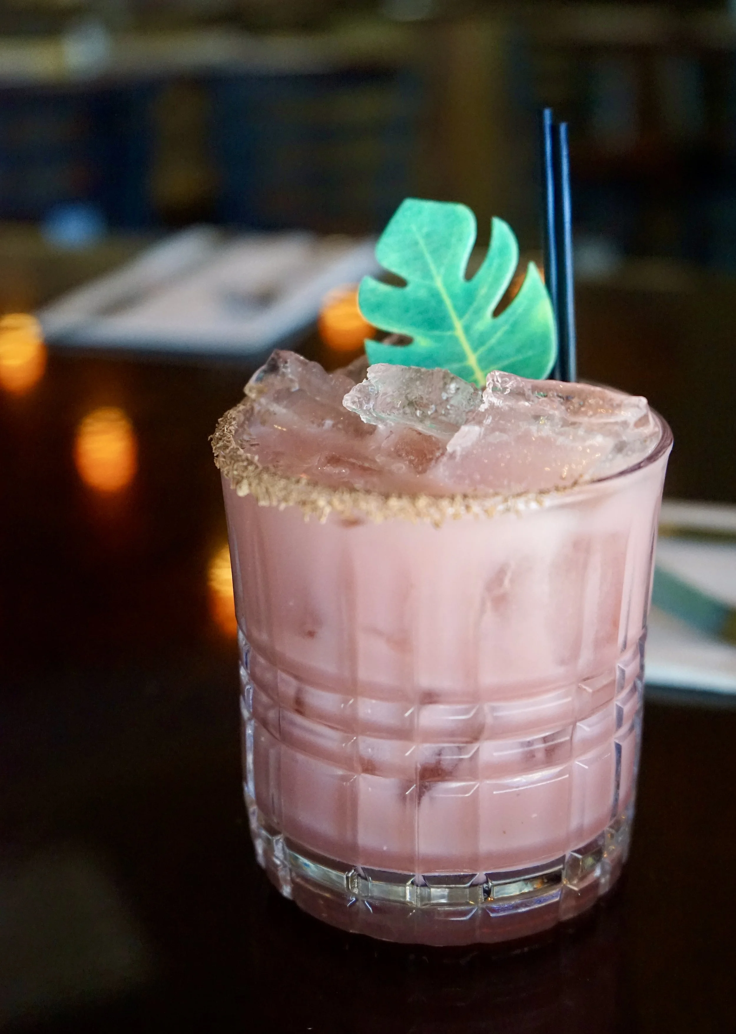 Pink cocktail in a glass with ice and a leaf garnish on a dark table.