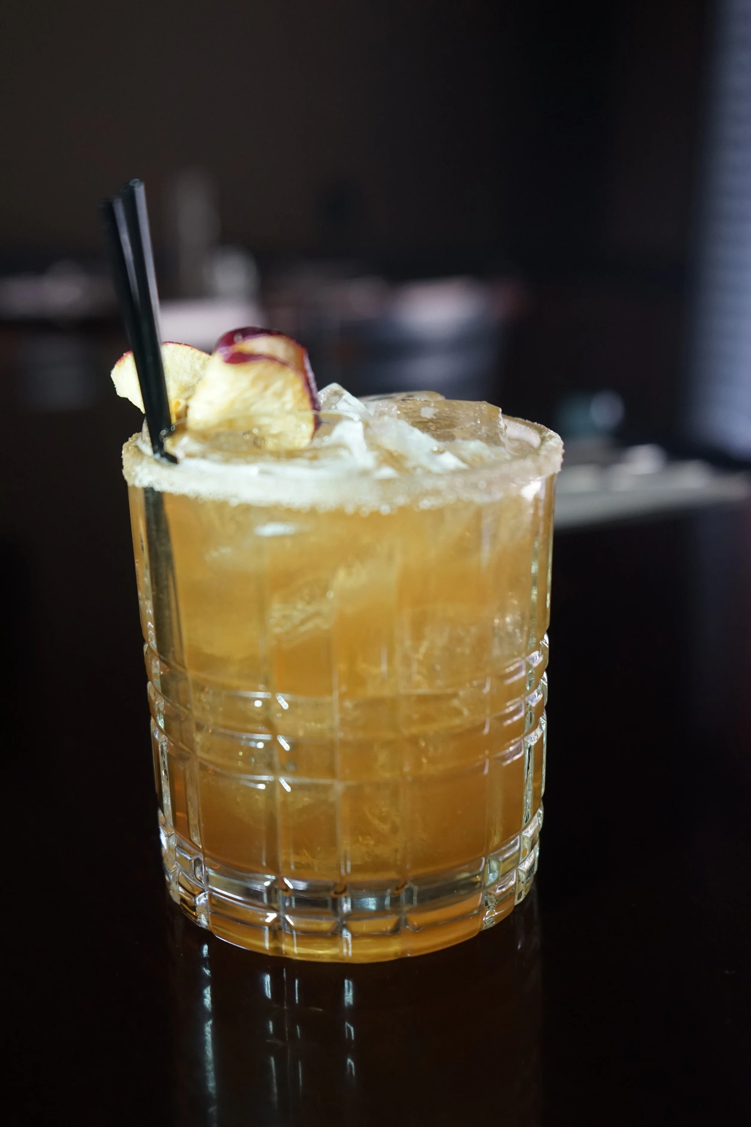 A glass of iced apple cocktail with black straws and apple garnish on a dark background.