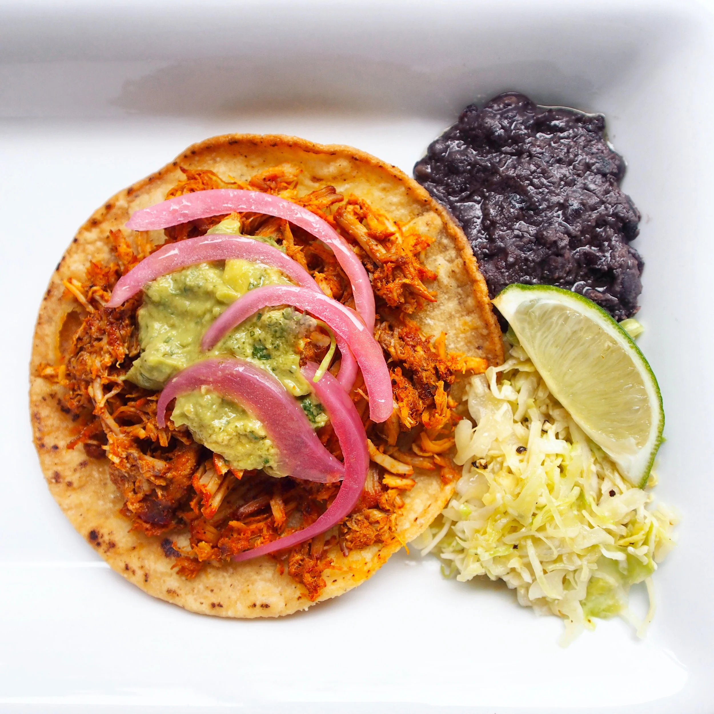 A cochinita pibil taco is served with barbecued pork, avocado salsa, and pickled red onions. Served on the side with cabbage slaw and refried beans! An easy taco recipe, but takes some time.