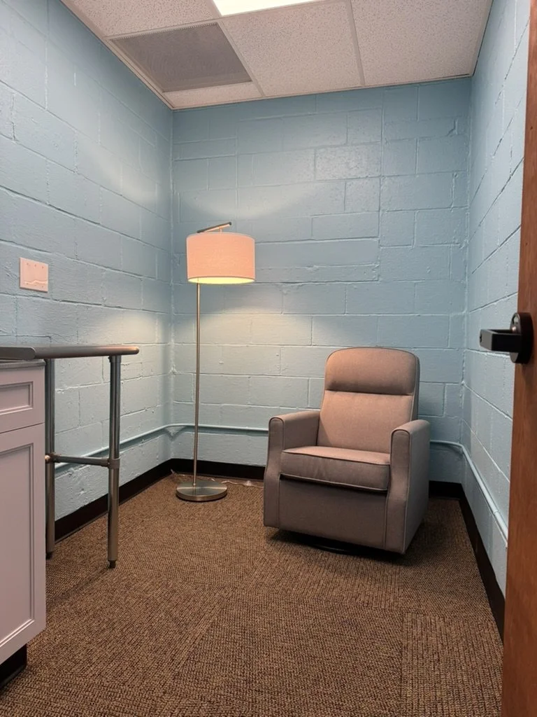 A small, plain room with light blue cinder block walls, a beige armchair, a floor lamp with a pink shade, a small white cabinet, and a brown carpeted floor.