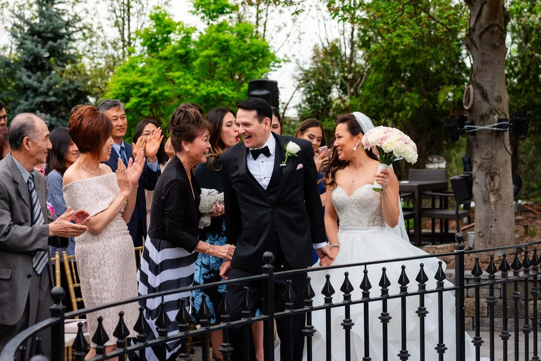 A wedding celebration with a bride and groom holding hands, surrounded by friends and family outdoors, with trees and greenery in the background.