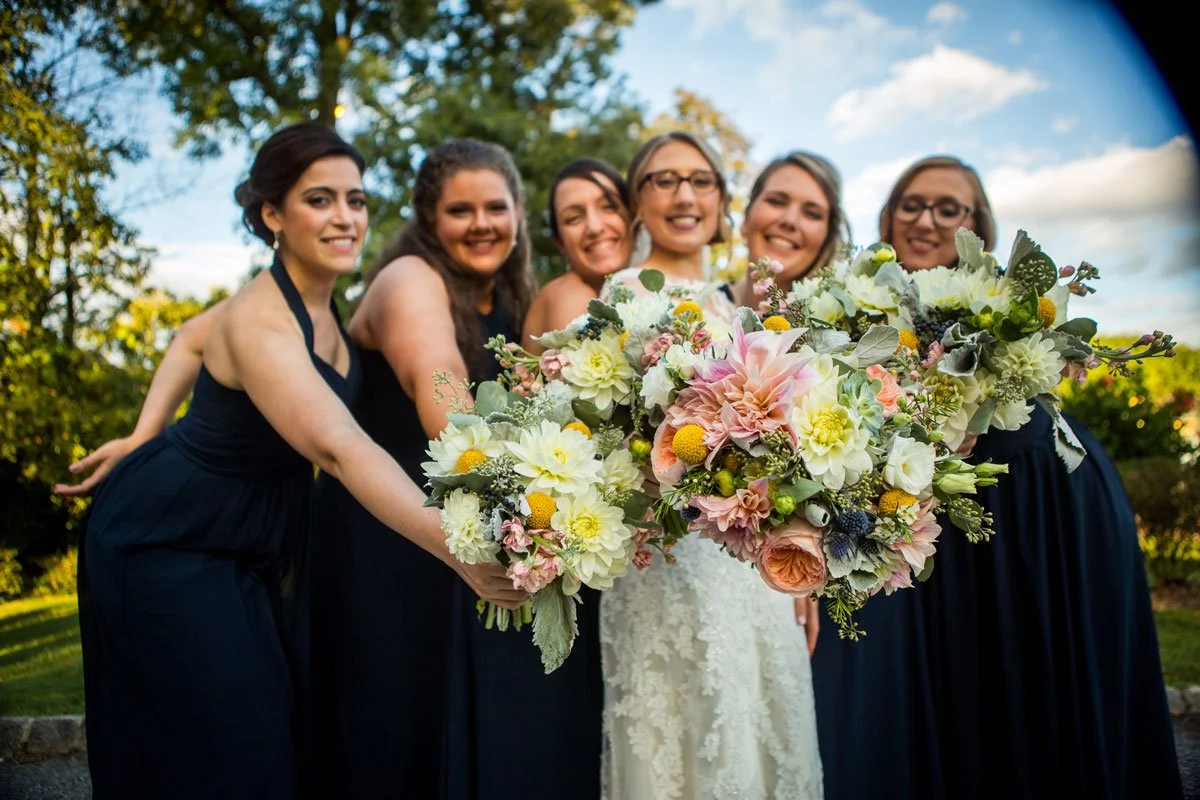 A group of six women in black dresses, one in white wedding dress, holding a large bouquet of flowers outdoors with trees and blue sky in the background.