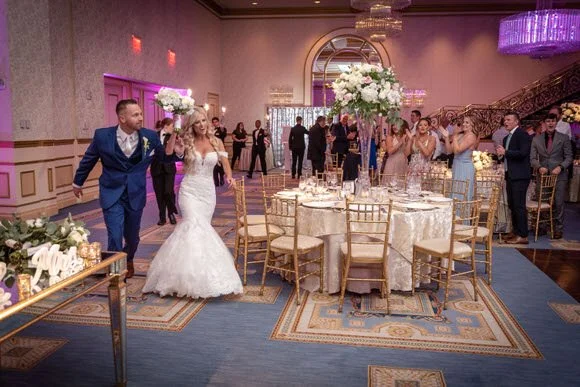 A bride and groom walking into a wedding reception hall, surrounded by guests clapping and taking photos. The hall has decorated tables, floral centerpieces, and elegant lighting.