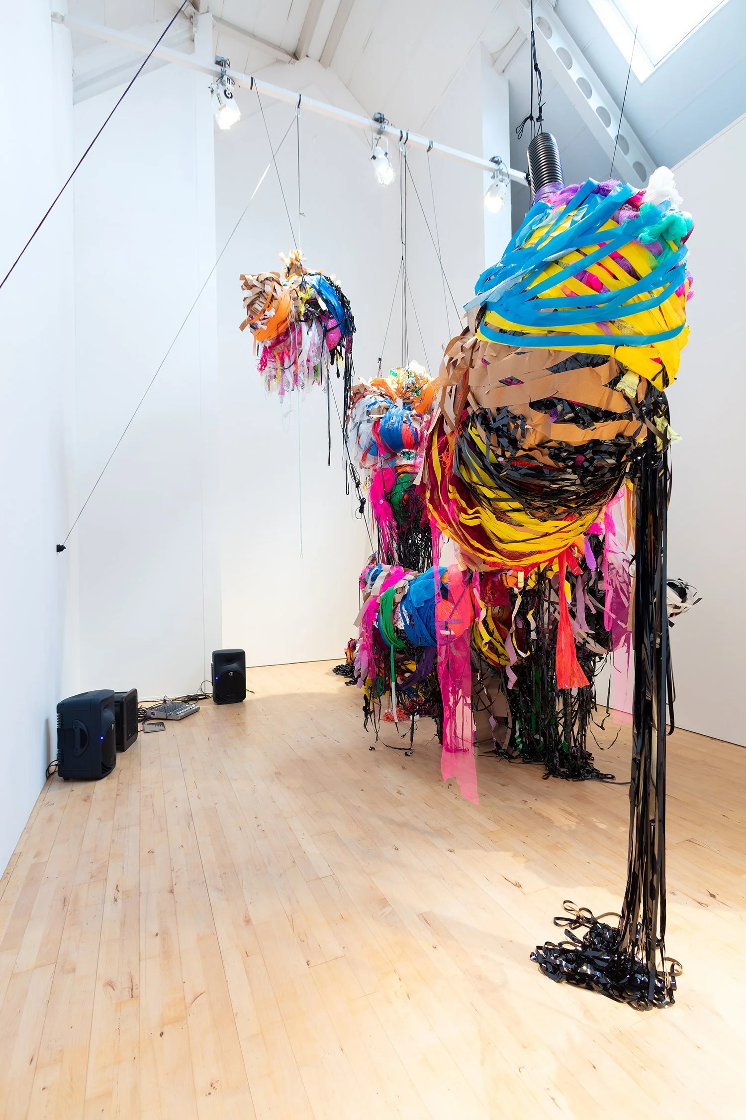 View along the installation with a prominent foreground sculpture wrapped in yellow, blue, and brown strips; additional suspended forms extend into the background.