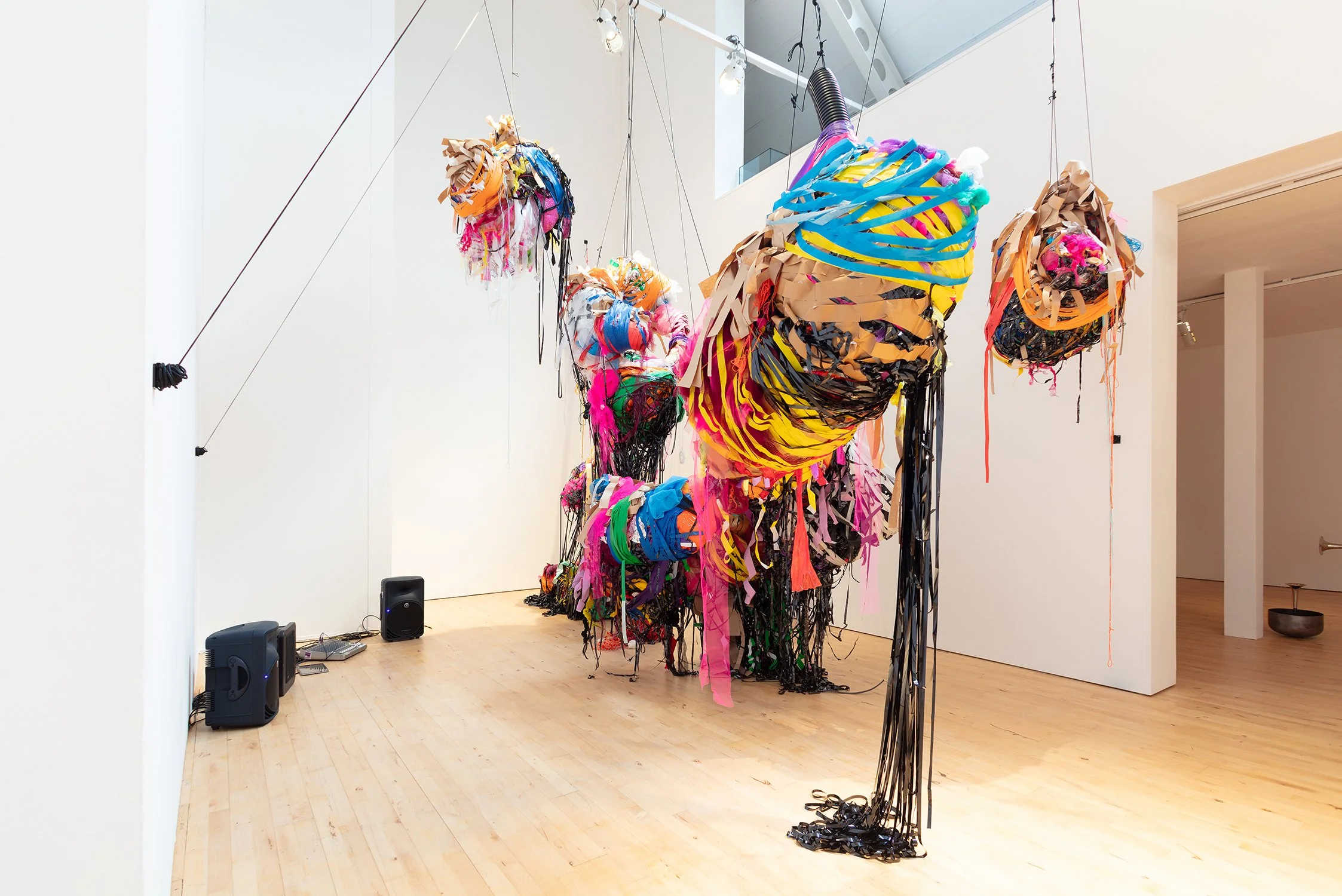 Installation view of multiple suspended, cocoon-like sculptures made from layered, brightly coloured materials, hanging at varying heights within a white gallery space.