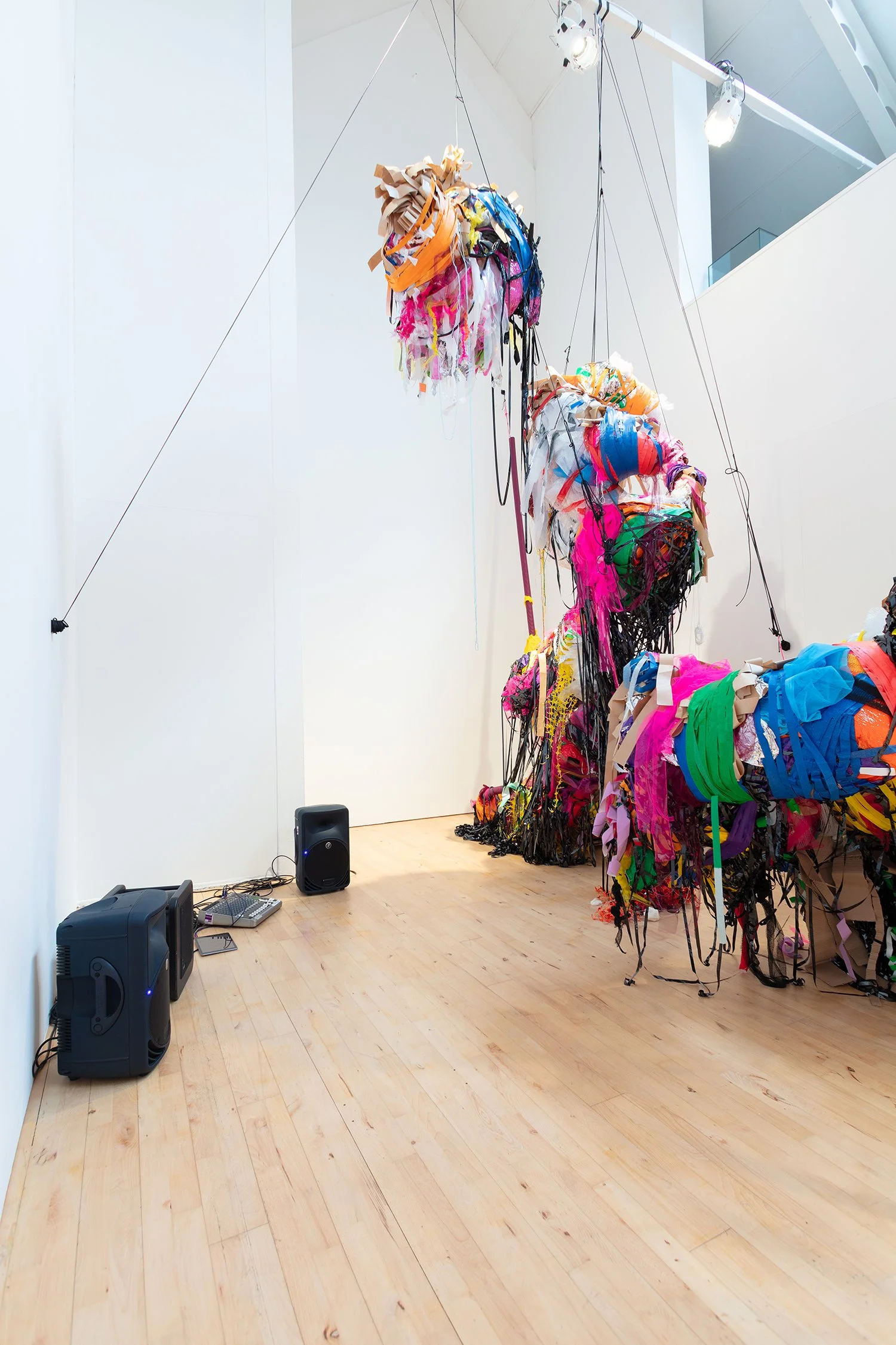Wide view of the installation showing multiple suspended sculptures arranged in a loose formation, with strong overhead lighting and exposed ceiling beams.
