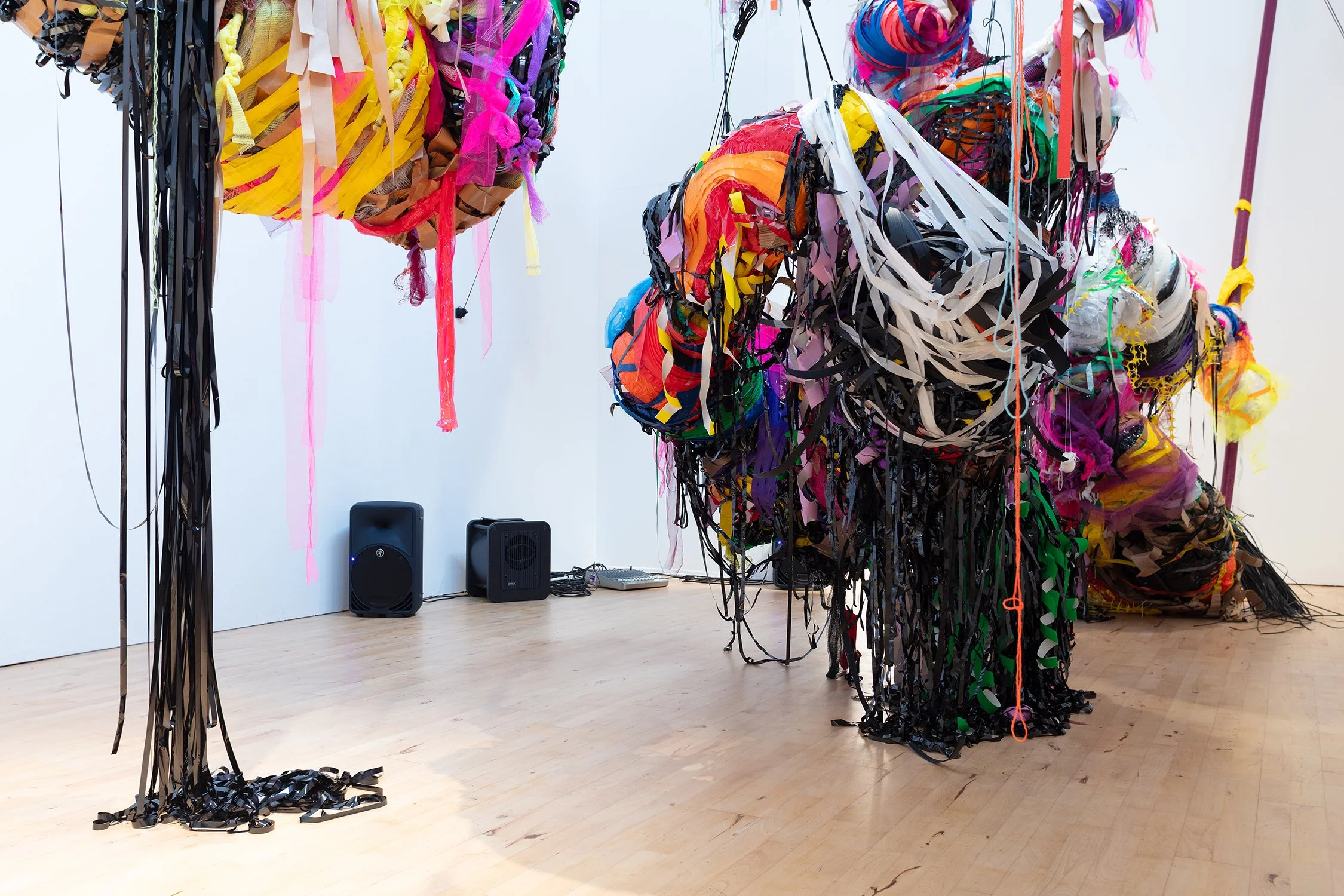 Low-angle view beneath the installation showing dense clusters of black tape and coloured materials trailing to the floor; speakers and cables sit against the wall behind.