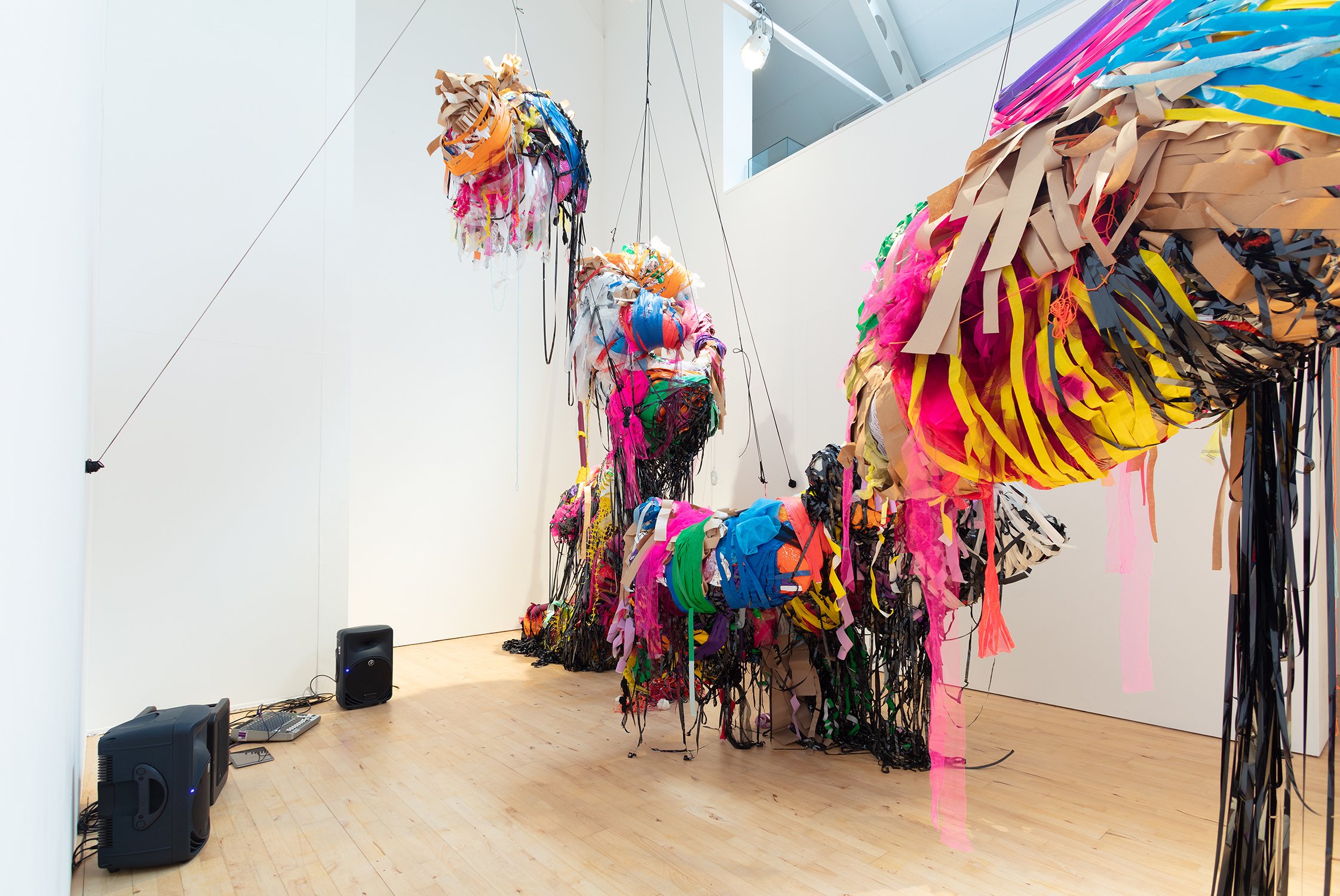 Side view of large, hanging sculptural forms composed of multicoloured strips of fabric and plastic, cascading downwards; speakers and audio equipment are visible on the floor nearby.