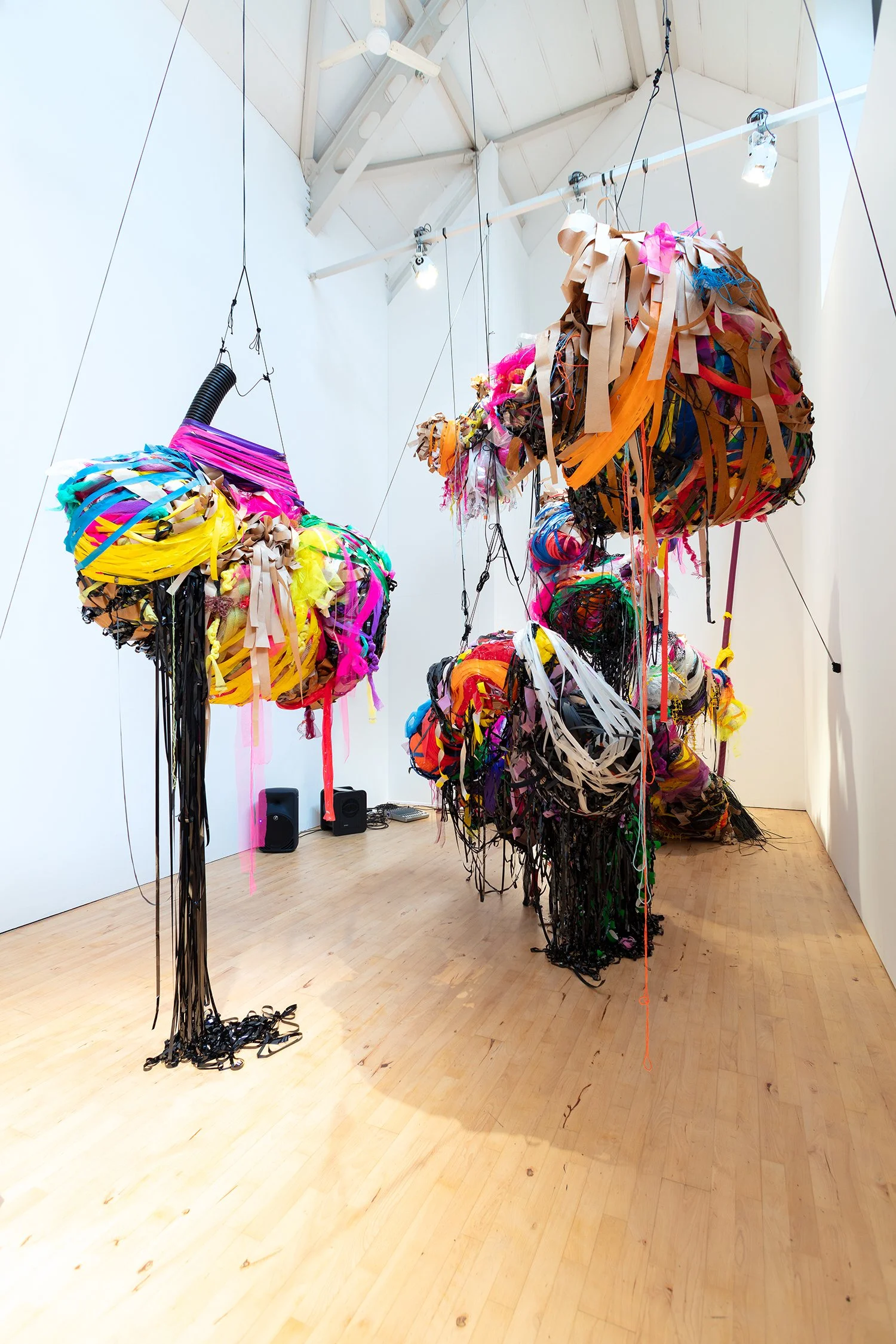 Vertical view of several suspended, bulbous sculptural forms wrapped in vivid strips of plastic and fabric, hanging in a tall, white gallery space.