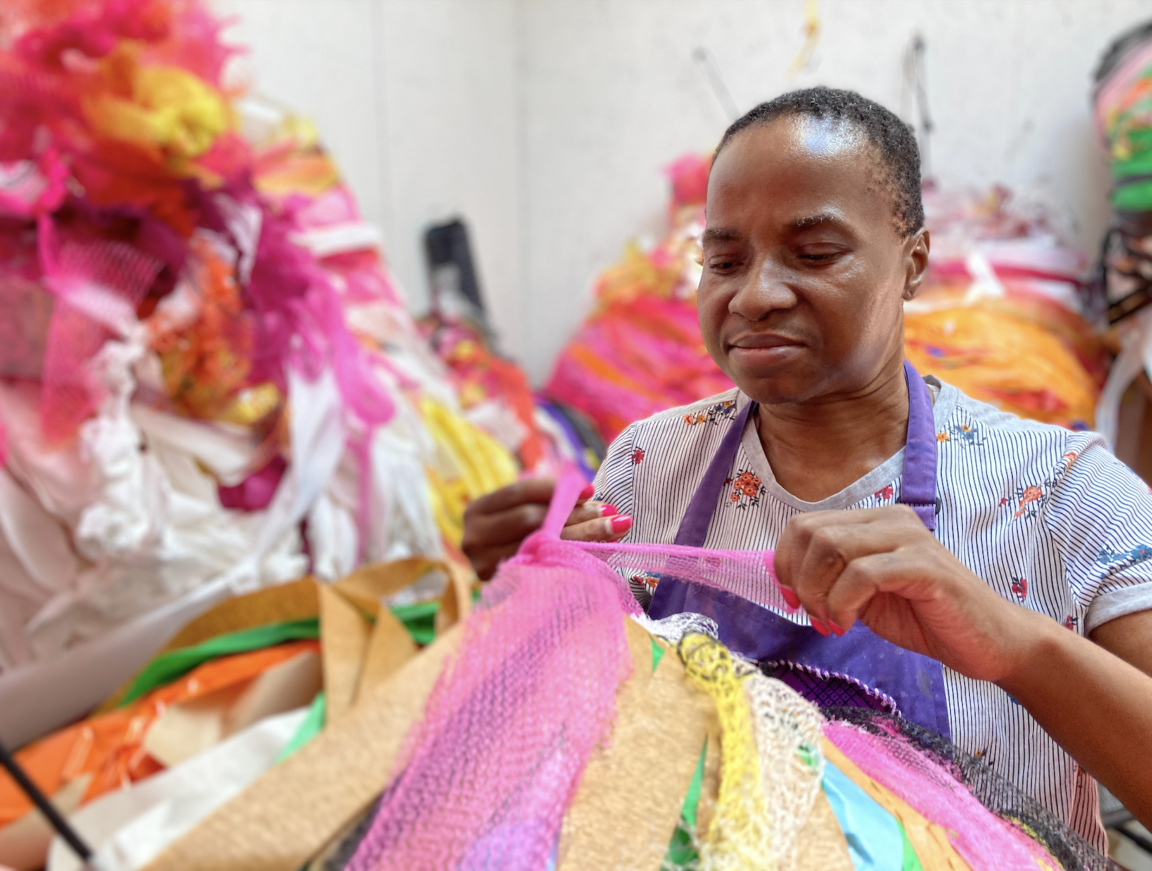 Artist Nnena Kalu in the studio surrounded by her colourful sculptures. She is braiding or tie-ing pink tulle on one of her sculptures. She has fuschia nail polish on.