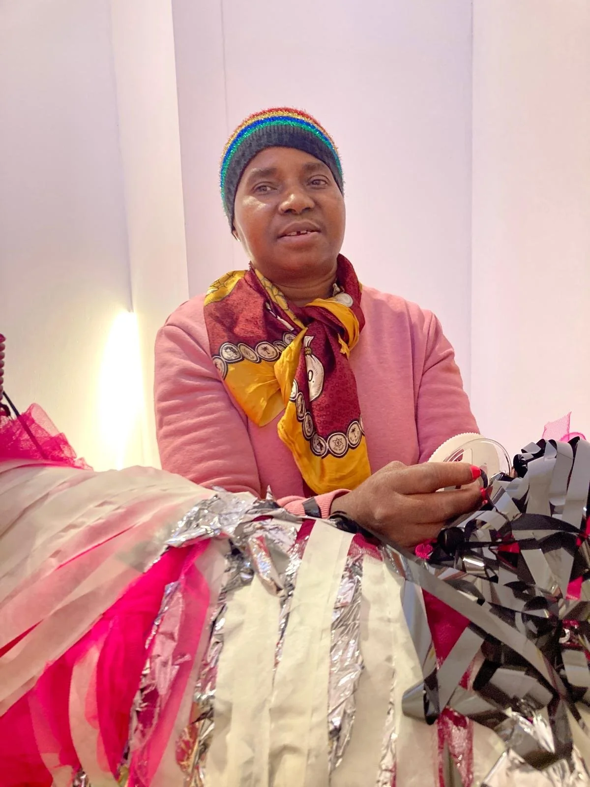 A woman wearing a rainbow-striped hat and scarf works with layered materials, holding tape and fabric elements as part of a large, colourful sculptural form.