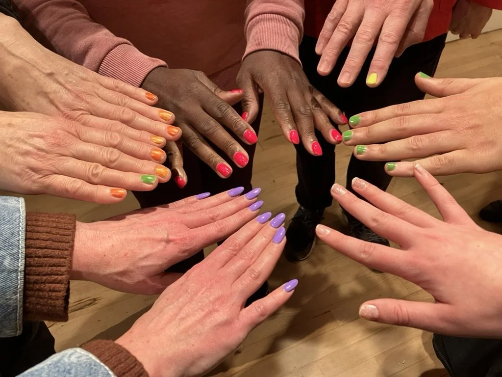 A group of people extend their hands in a circle, each with brightly painted nails in different colours, creating a shared, playful composition.