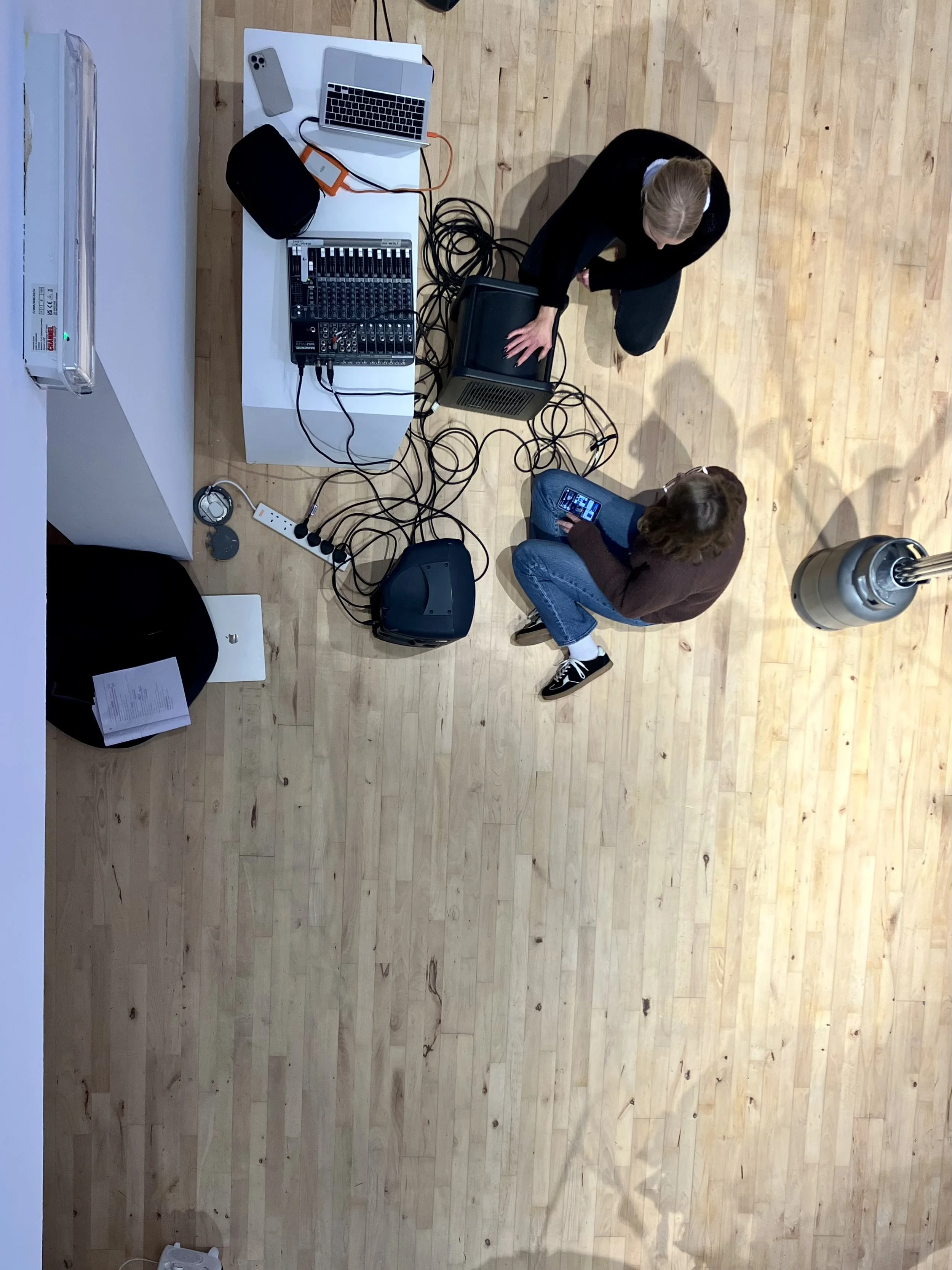 Overhead view of two people seated on the floor working with audio equipment, cables, and a laptop arranged on a table in a gallery space.